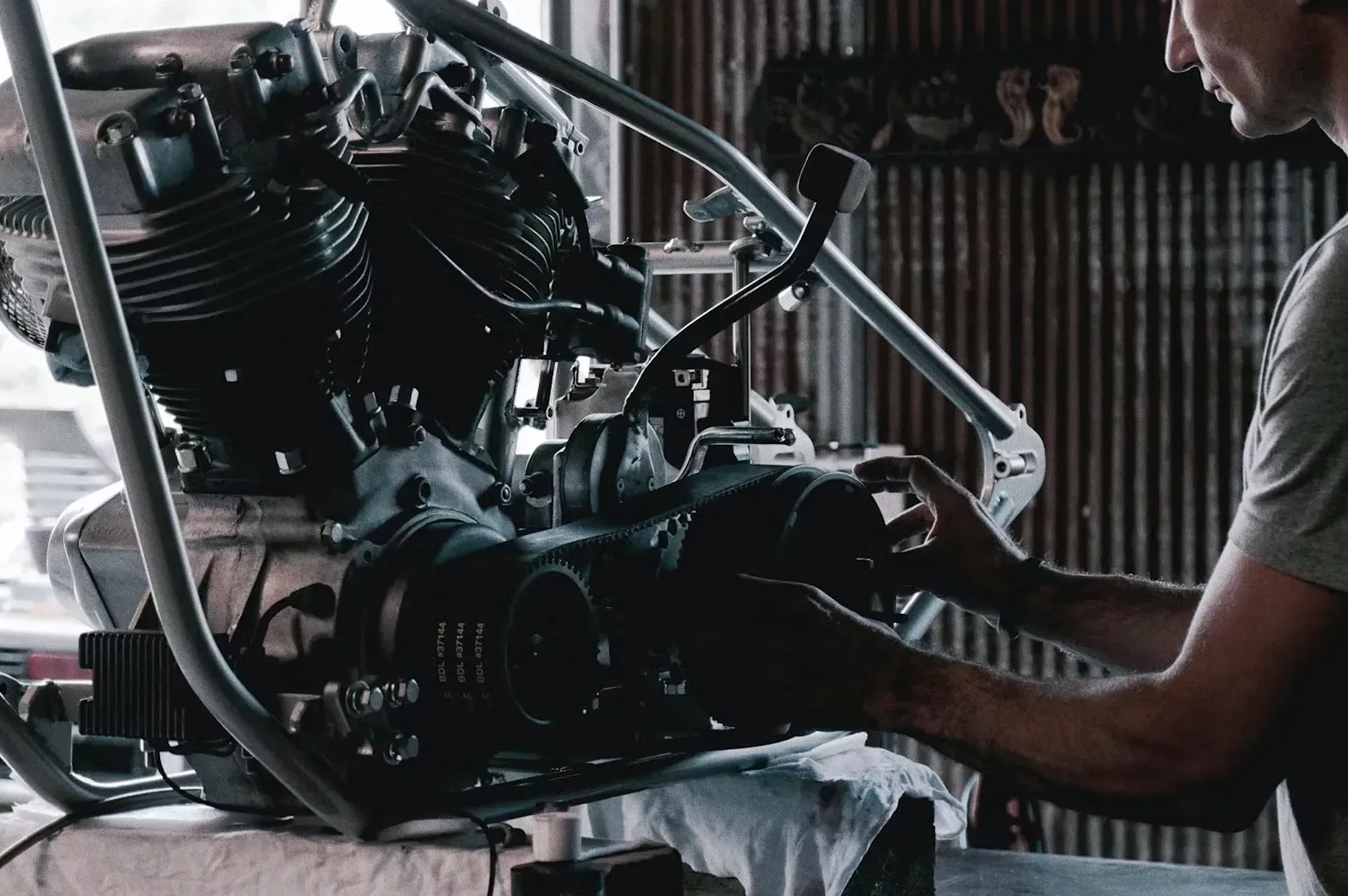 Three‑quarter side angle close‑up shot of a motorcycle engine mounted on a workbench, with a person’s hands adjusting mechanical components inside a workshop setting.
