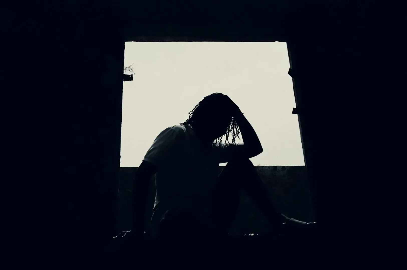 Wide shot silhouette of a person sitting inside a dark doorway, head resting on hand, backlit by daylight, conveying loneliness, stress, and emotional struggle.