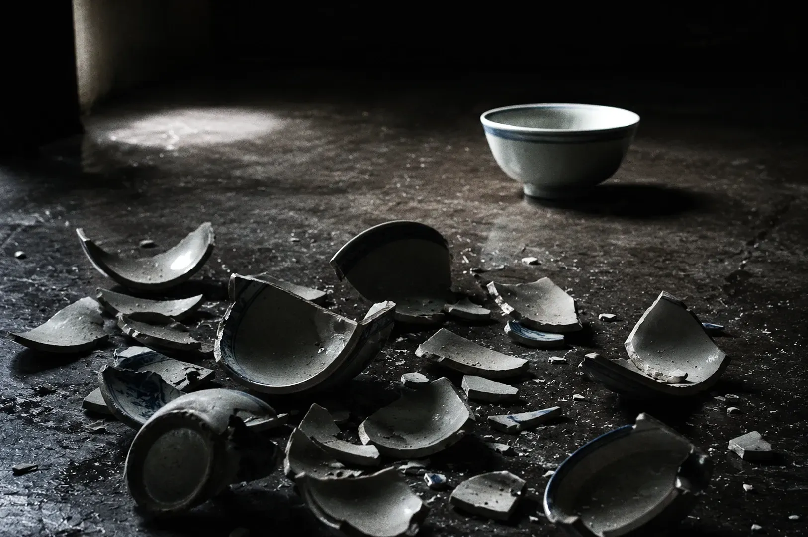 High-angle shot of shattered ceramic bowls on dark surface with intact bowl in background representing waste, perfection, and hidden costs of craftsmanship