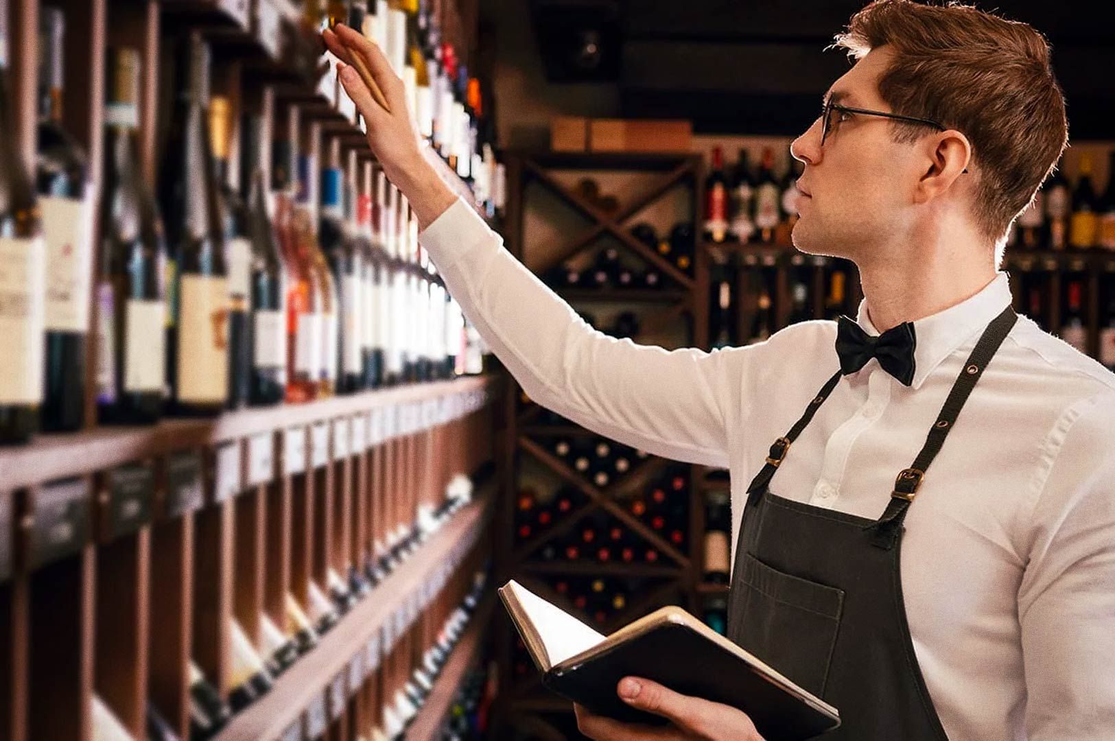 Professional sommelier in bow tie and apron examining wine bottle in cellar, notebook in hand.