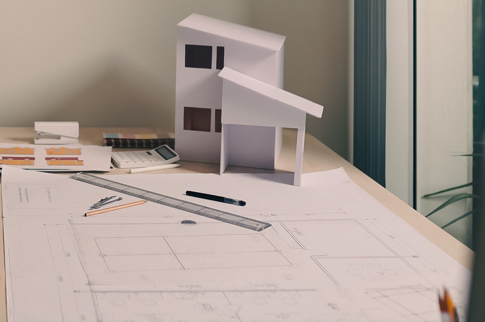 Architectural workspace with house models, blueprints, and drafting tools on a desk near a window.