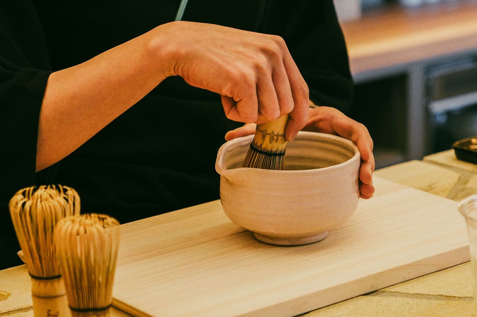 Hands whisking frothy matcha in a ceramic bowl with bamboo chasen.