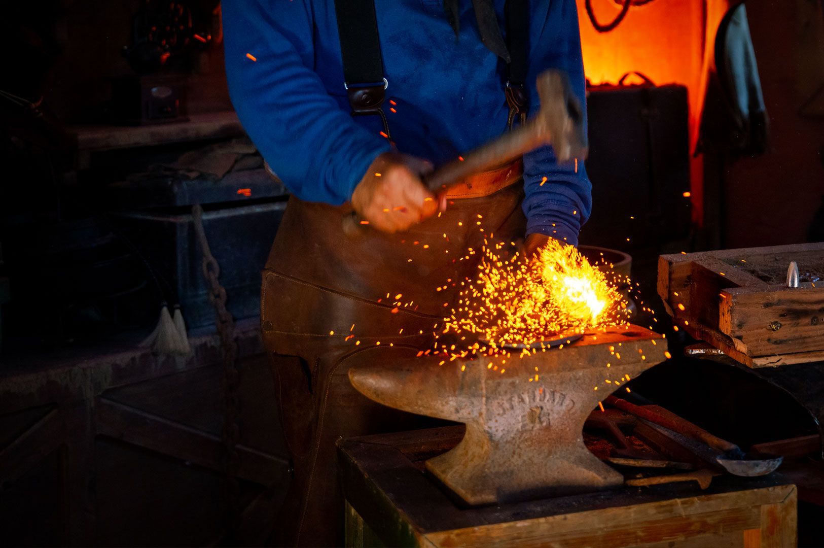 Blacksmith hammering heated metal on an anvil with sparks flying inside a dimly lit workshop.