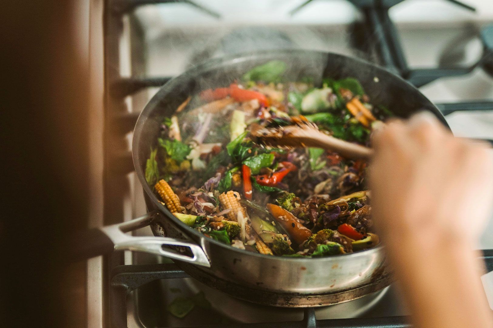Colorful mix of vegetables being stir‑fried in a steaming pan on a stovetop with a wooden spatula.