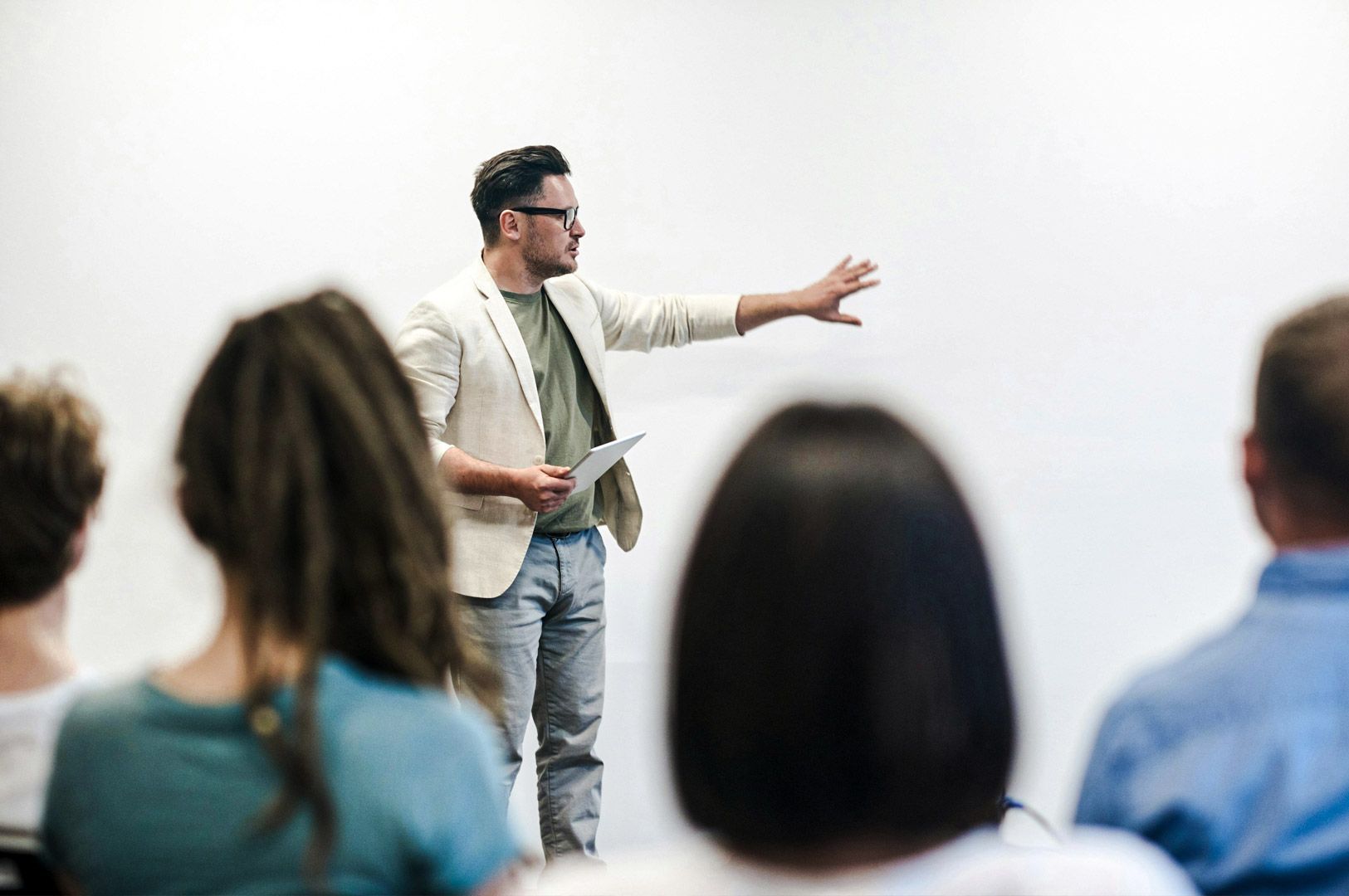 A speaker standing in front of an audience while presenting with one arm extended toward a blank wall, holding notes in the other hand.
