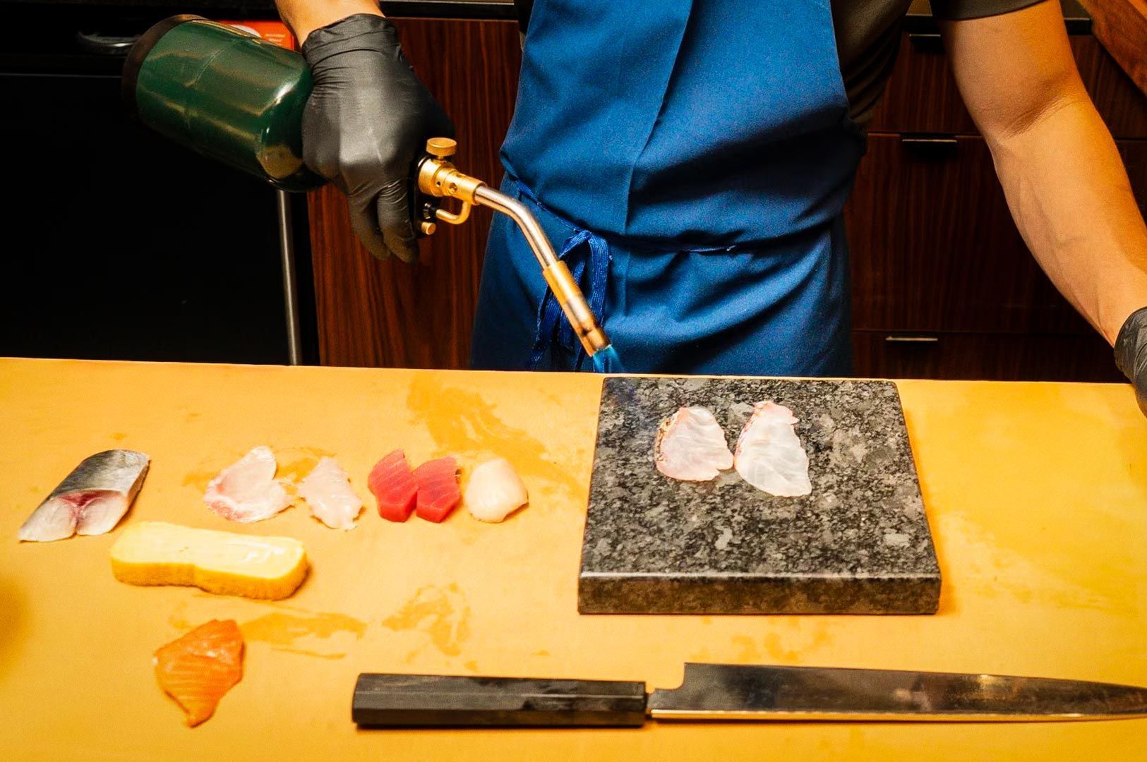 A person wearing gloves and an apron using a handheld torch to sear pieces of fish laid out on a cutting board, with assorted sushi ingredients and a large knife nearby.