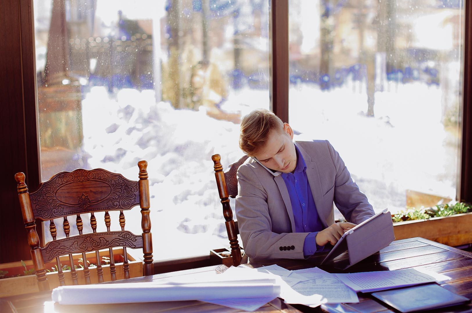 Professional working on a tablet at a wooden desk with documents spread out and large windows showing a snowy outdoor landscape.