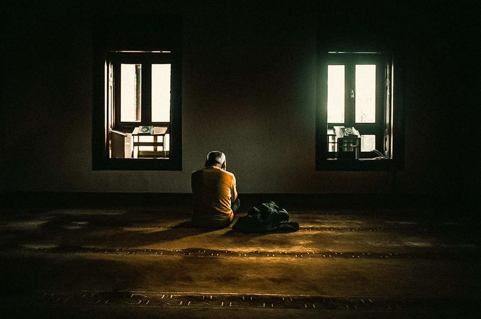 Person sitting on the floor of a dimly lit room between two windows, creating a peaceful and contemplative atmosphere.