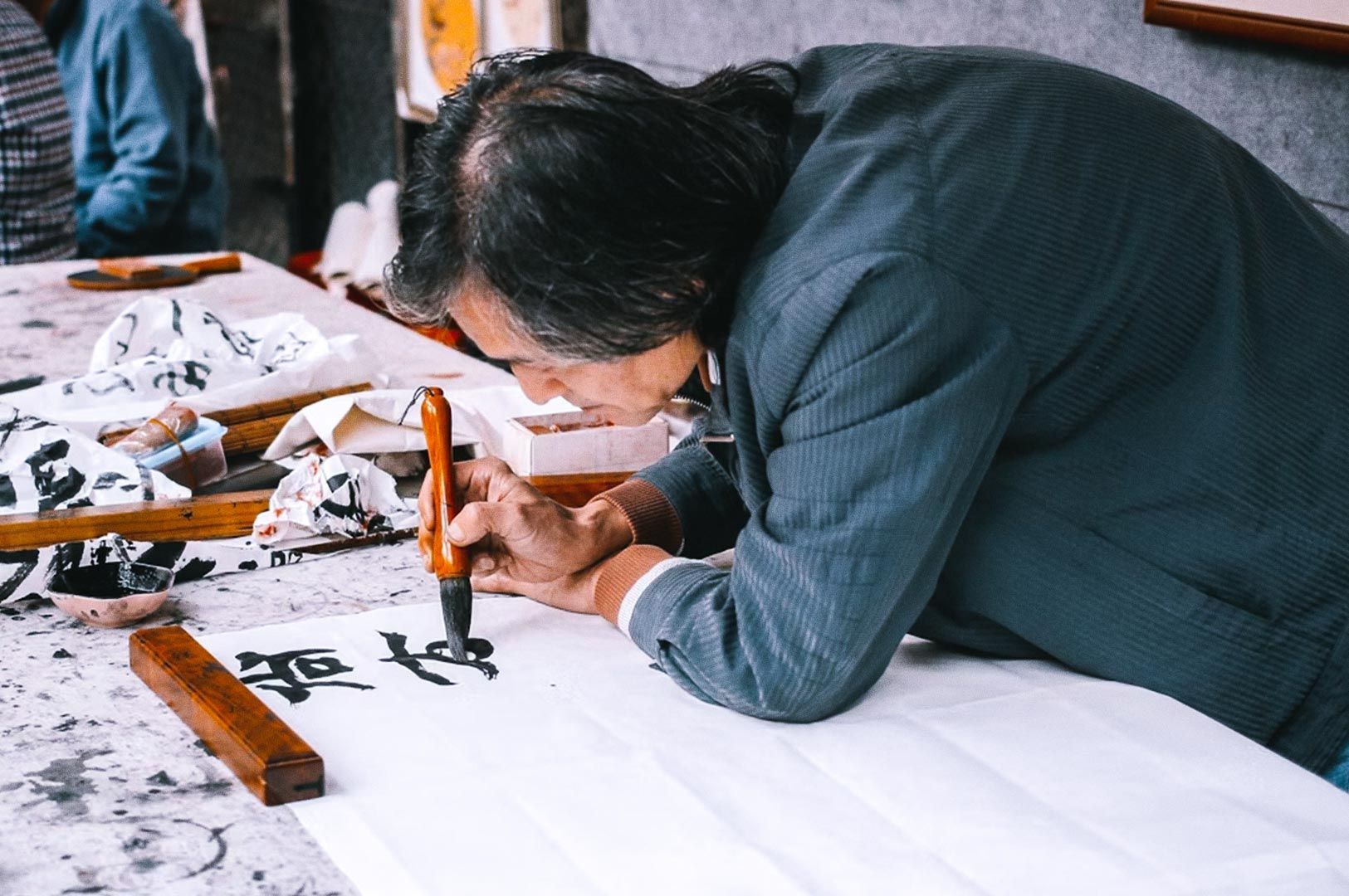 Person practicing traditional calligraphy, using a brush to paint black ink characters on white paper at a worktable.