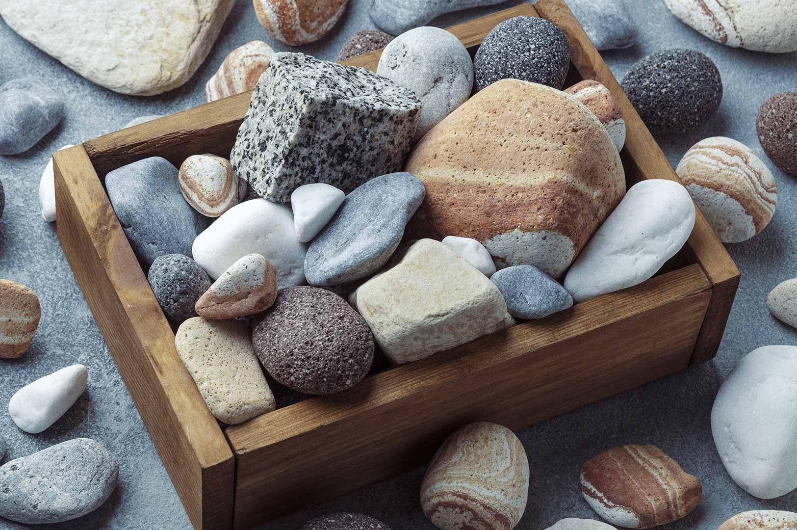 Wooden box filled with smooth, rounded stones and pebbles in white, gray, black, brown, and beige, some showing layered or speckled patterns; extra stones scattered around the box and a distinctive speckled cube-shaped granite rock visible.