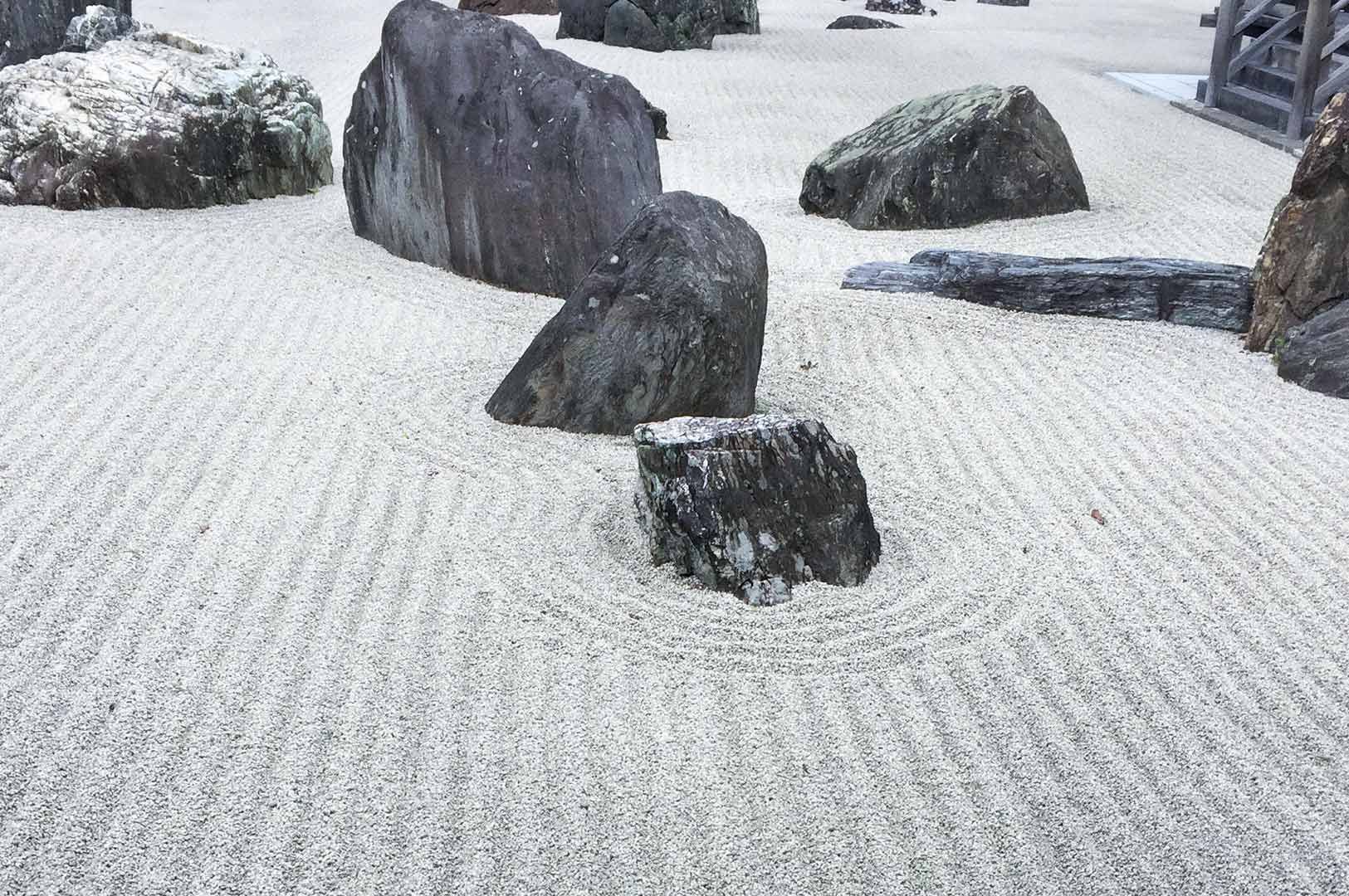 Japanese Zen rock garden with raked white gravel forming circular patterns around large natural stones, creating a minimalist and tranquil landscape scene.