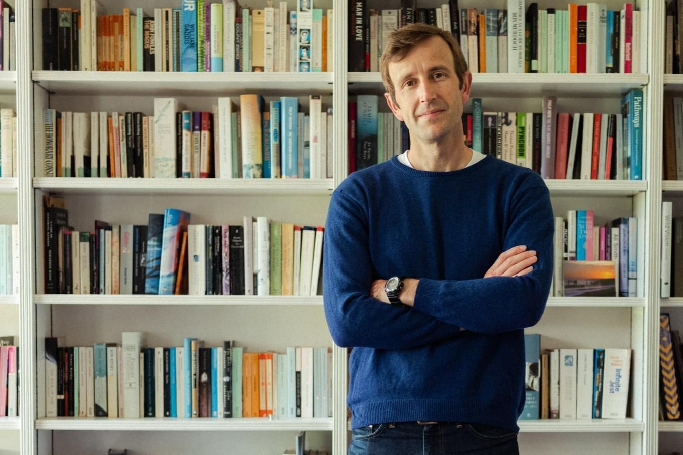 A man in a blue sweater stands with his arms crossed in front of white floor-to-ceiling bookshelves that are filled with various books. The collection behind him includes a wide range of colorful spines, suggesting a diverse and extensive library.