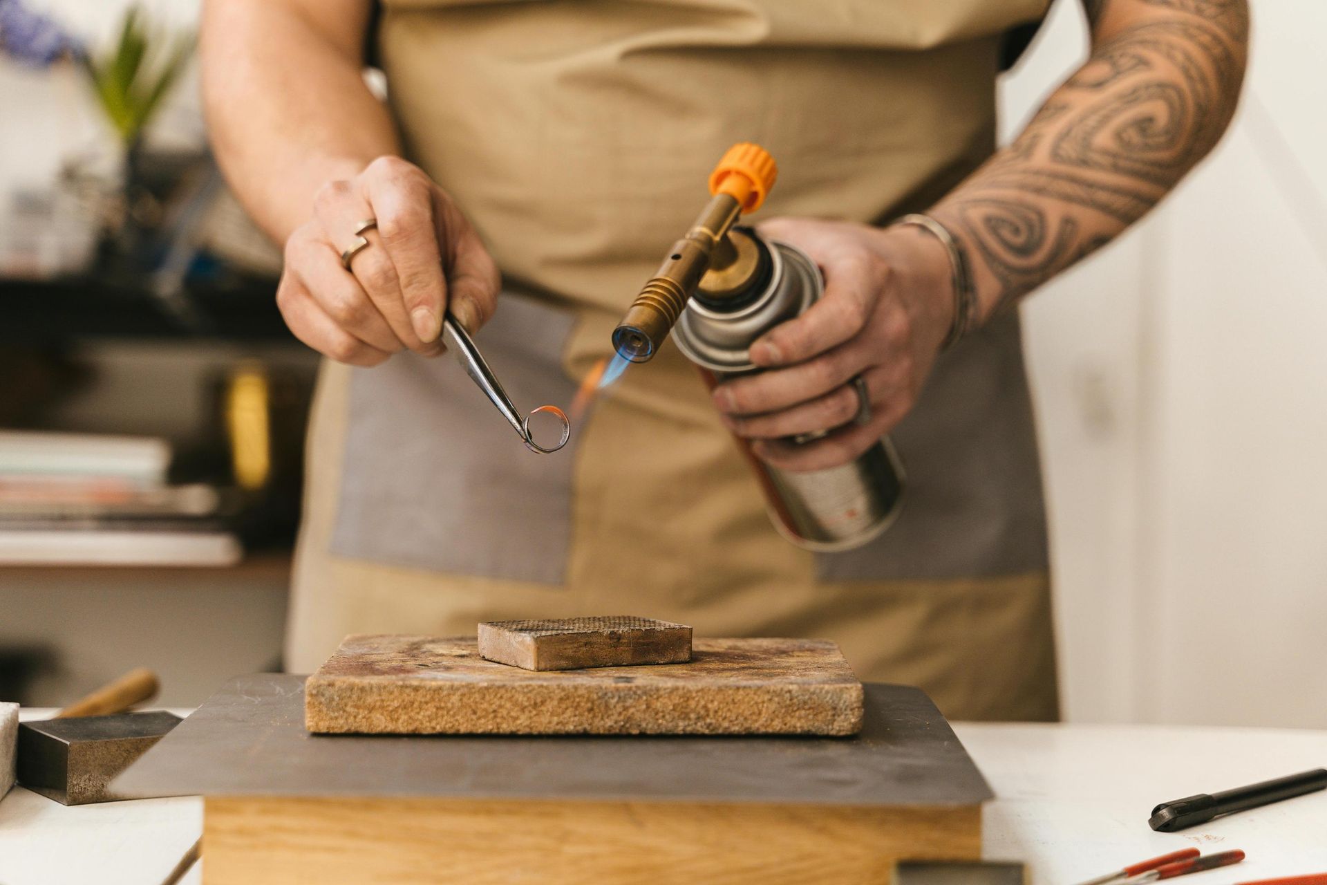 A person with a tattooed arm uses a handheld torch to apply heat to a small ring held with tweezers. They are wearing a tan apron and working over a heat-resistant block in a craft studio or workshop setting.