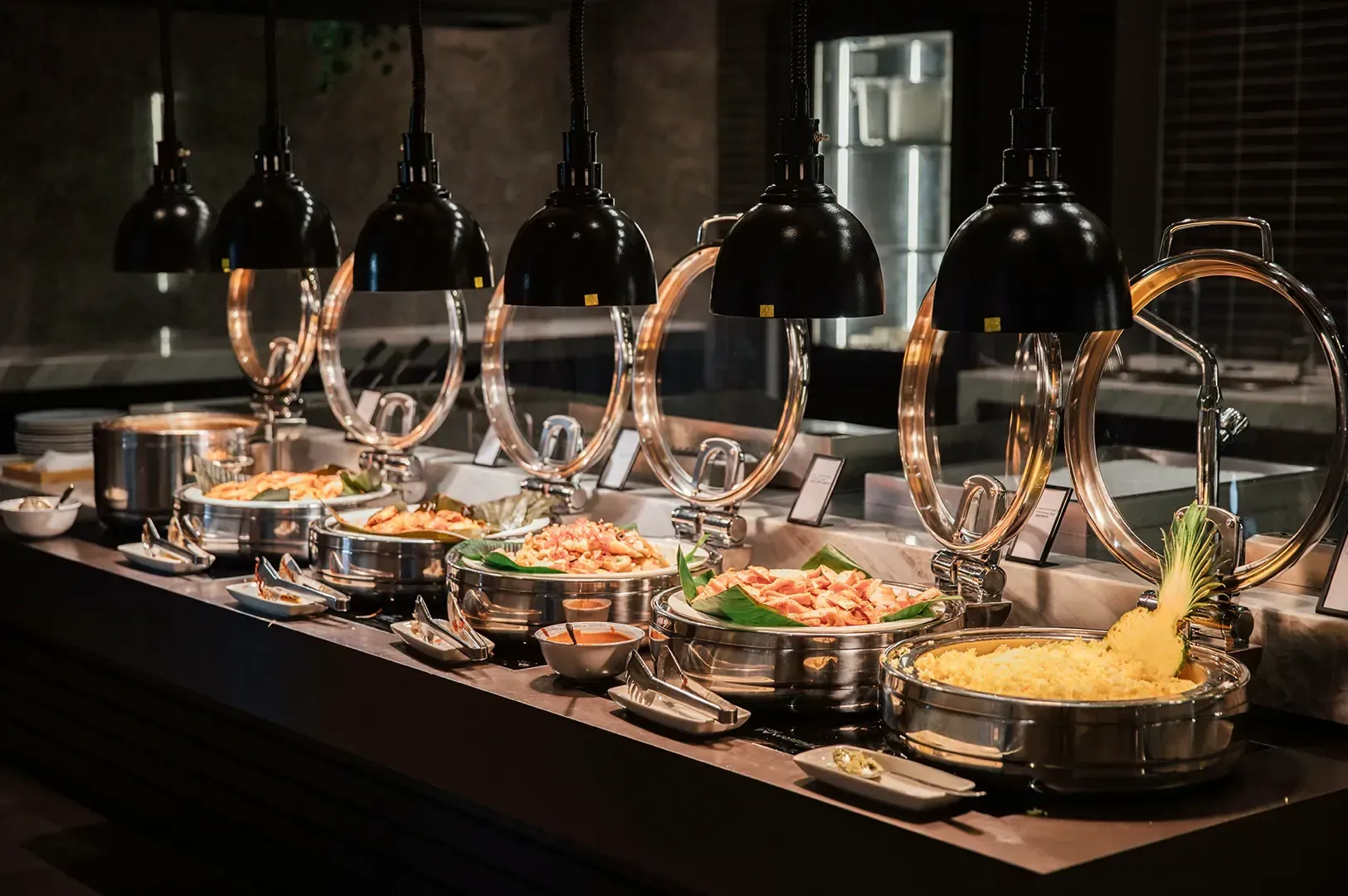 A line of stainless steel buffet food warmers filled with diverse dishes under hanging heat lamps, highlighting the wide range of culinary choices.