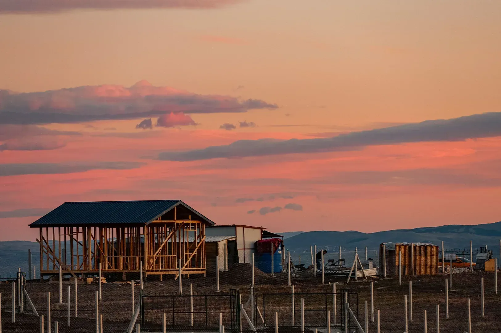 A wooden building frame under a vibrant, long-exposure sunset, illustrating the quiet transition of construction and the dedication required to create a lasting legacy for those we will never meet.