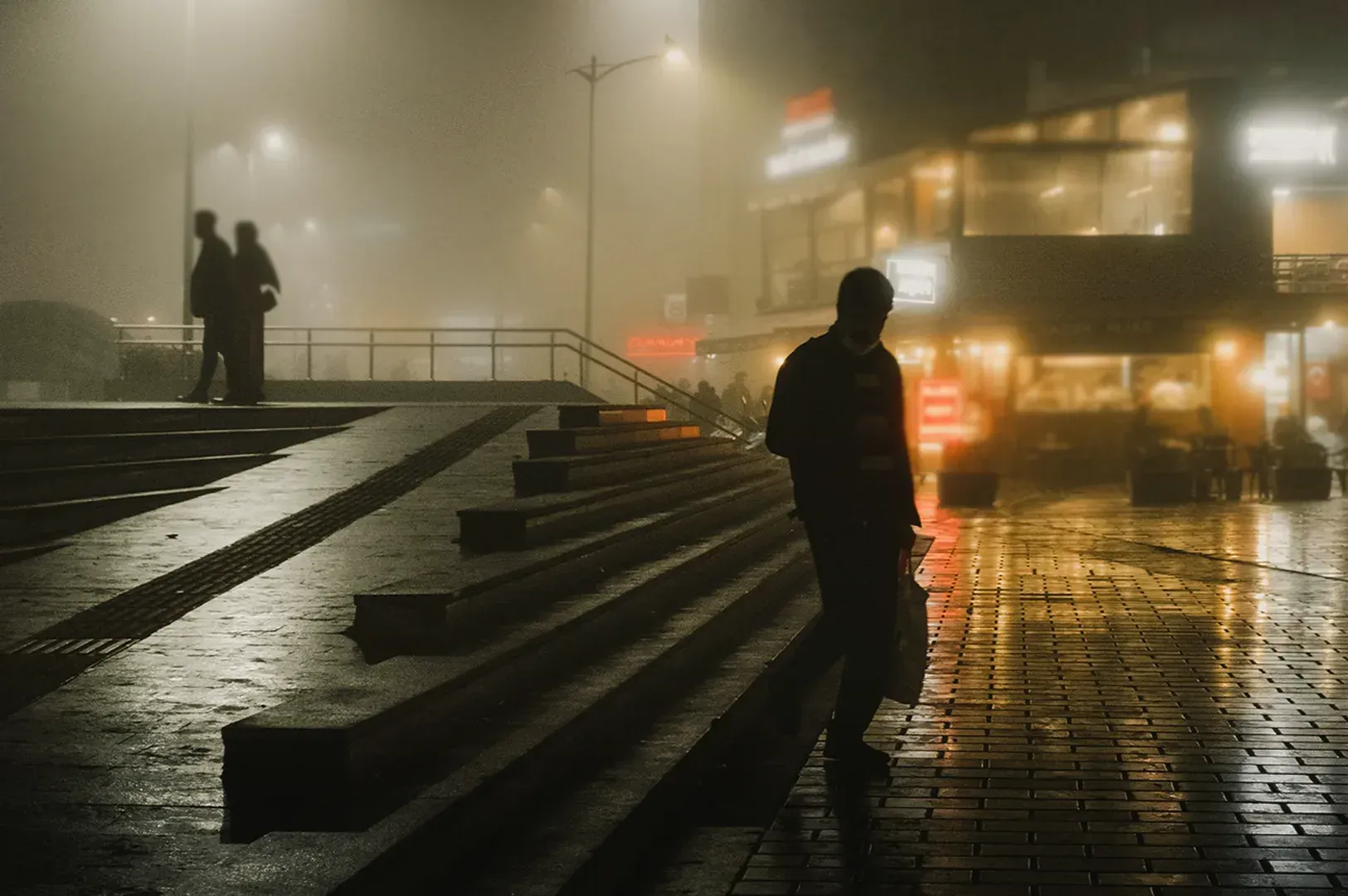 A lone figure walks through a foggy, dimly lit city square at night, reflecting on the quiet, solitary moments that bridge our human connections in the hospitality of strangers.
