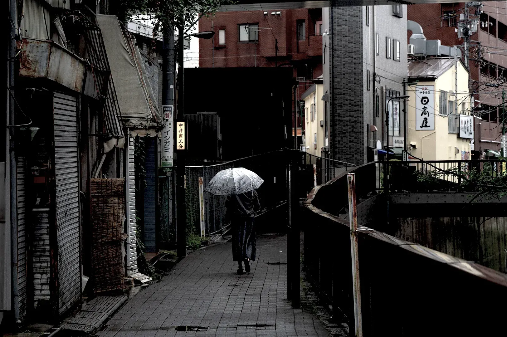 A solitary figure walking away down a narrow, quiet urban path under a clear umbrella, reflecting the theme of disciplined attention and wandering through city streets.