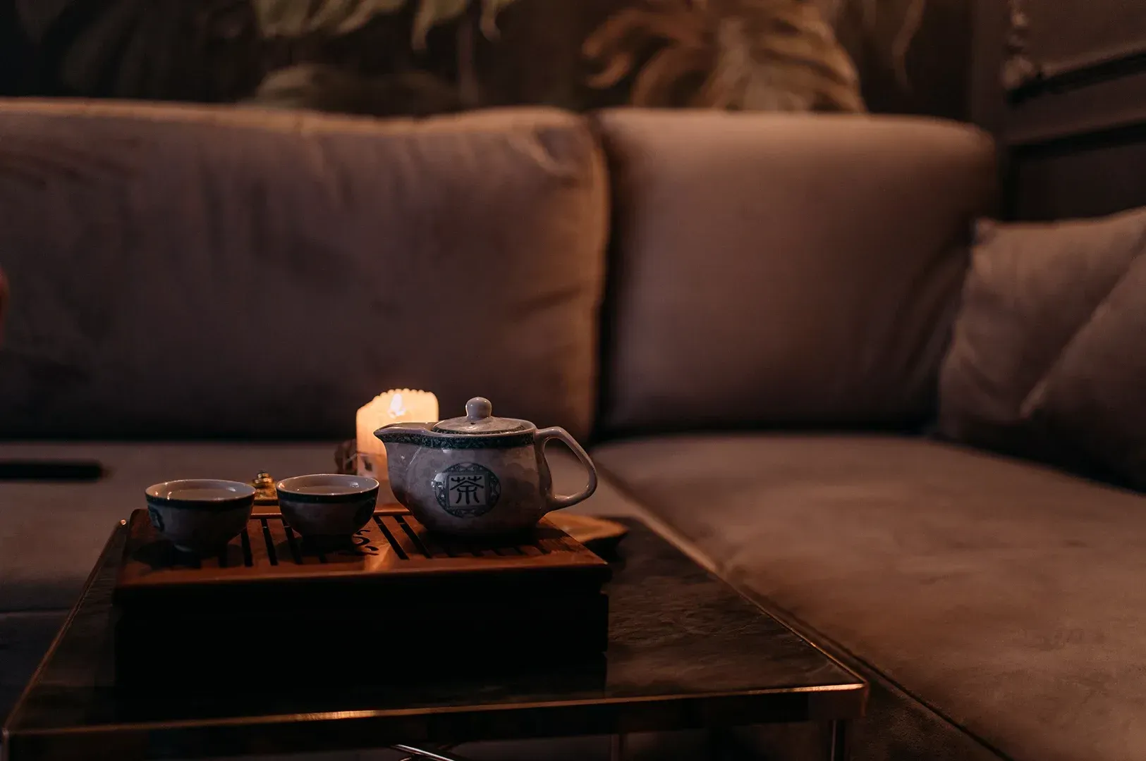 A peaceful, dimly lit tea room with a soft-glowing candle beside a teapot and cups on a wooden tray, set against a cozy sofa to invite the practice of listening when we stop performing.