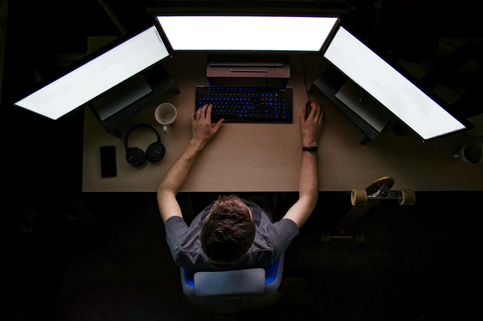 Overhead bird’s-eye view of a person working alone at a multi-monitor desk in a dark room, illustrating deep work, focus, and the modern struggle with digital distraction and solitude