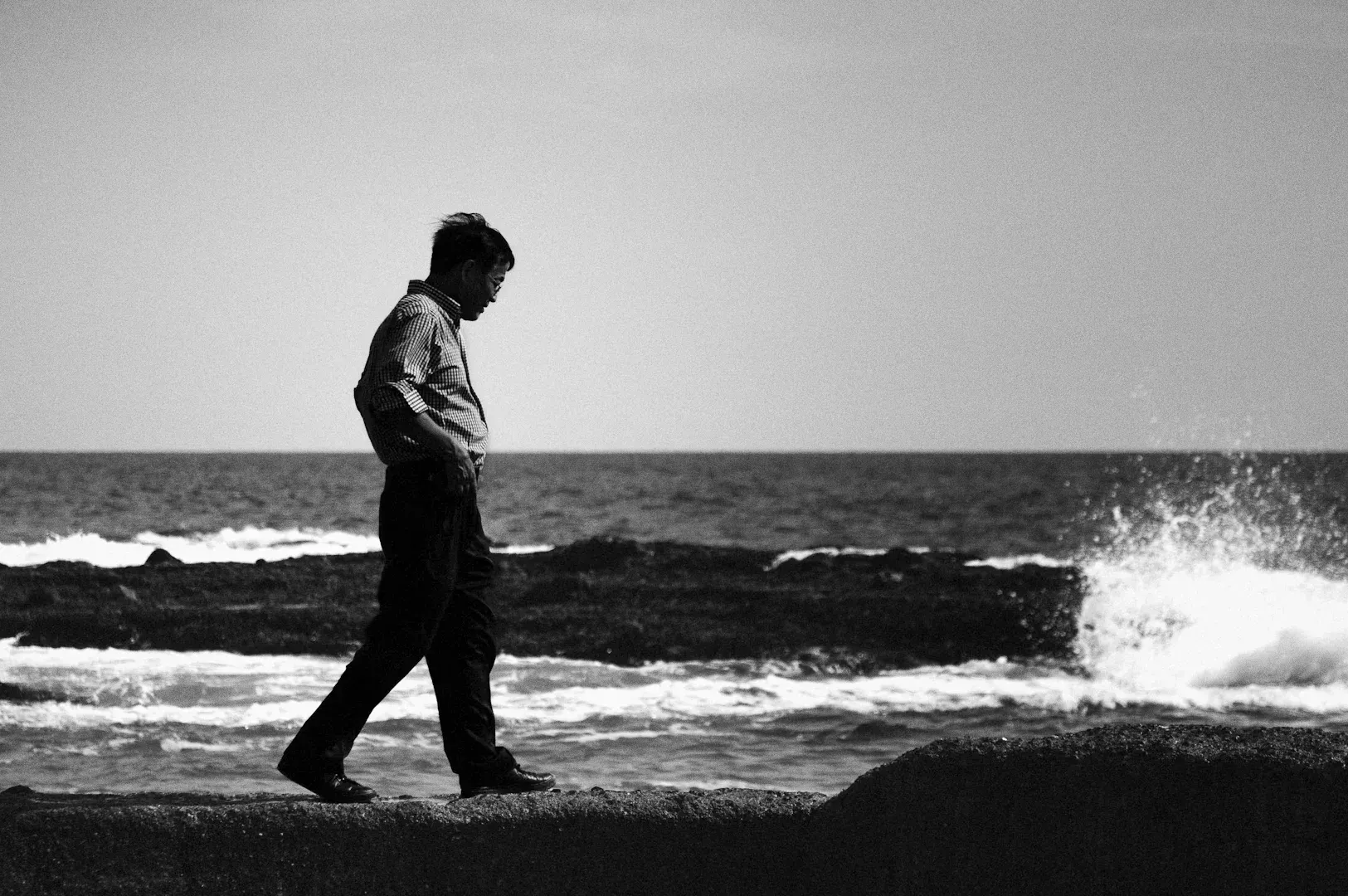 Wide-angle black-and-white photo of a man walking alone along a rocky shoreline, waves crashing nearby, captured from a side profile to evoke solitude, reflection, and personal journey.