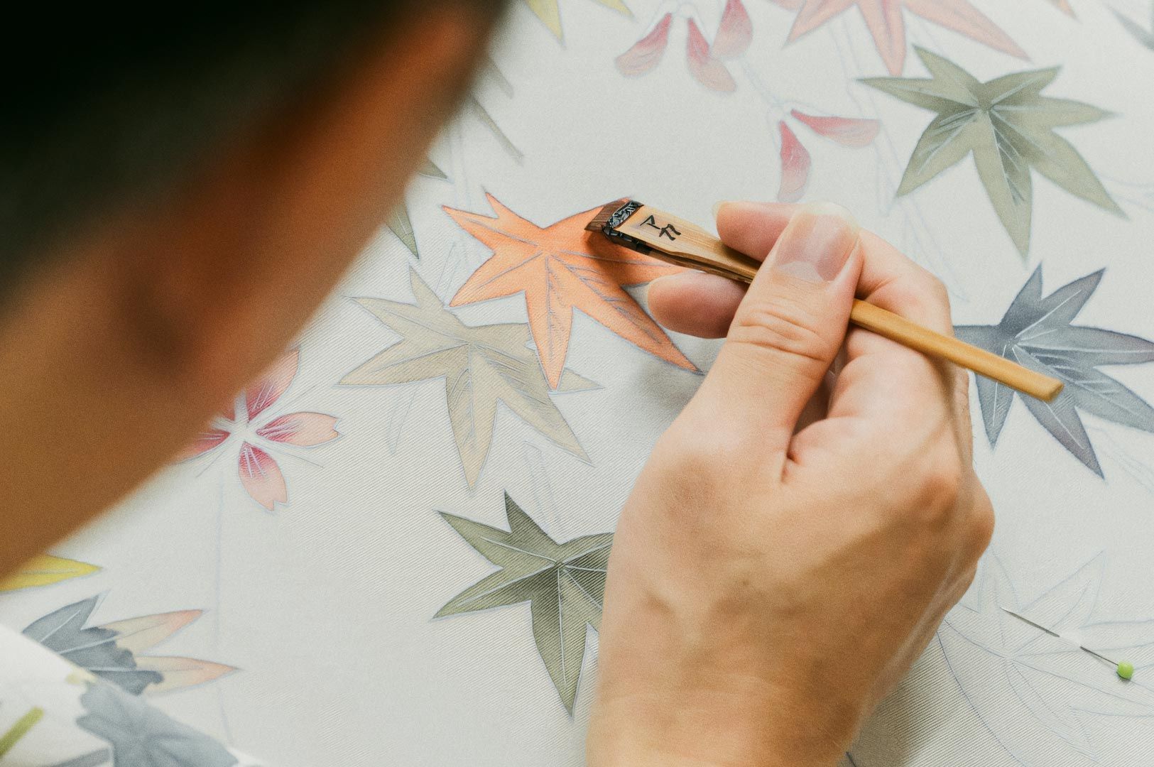 Artisan painting orange autumn-leaf pattern on fabric using a traditional Japanese dyeing tool.