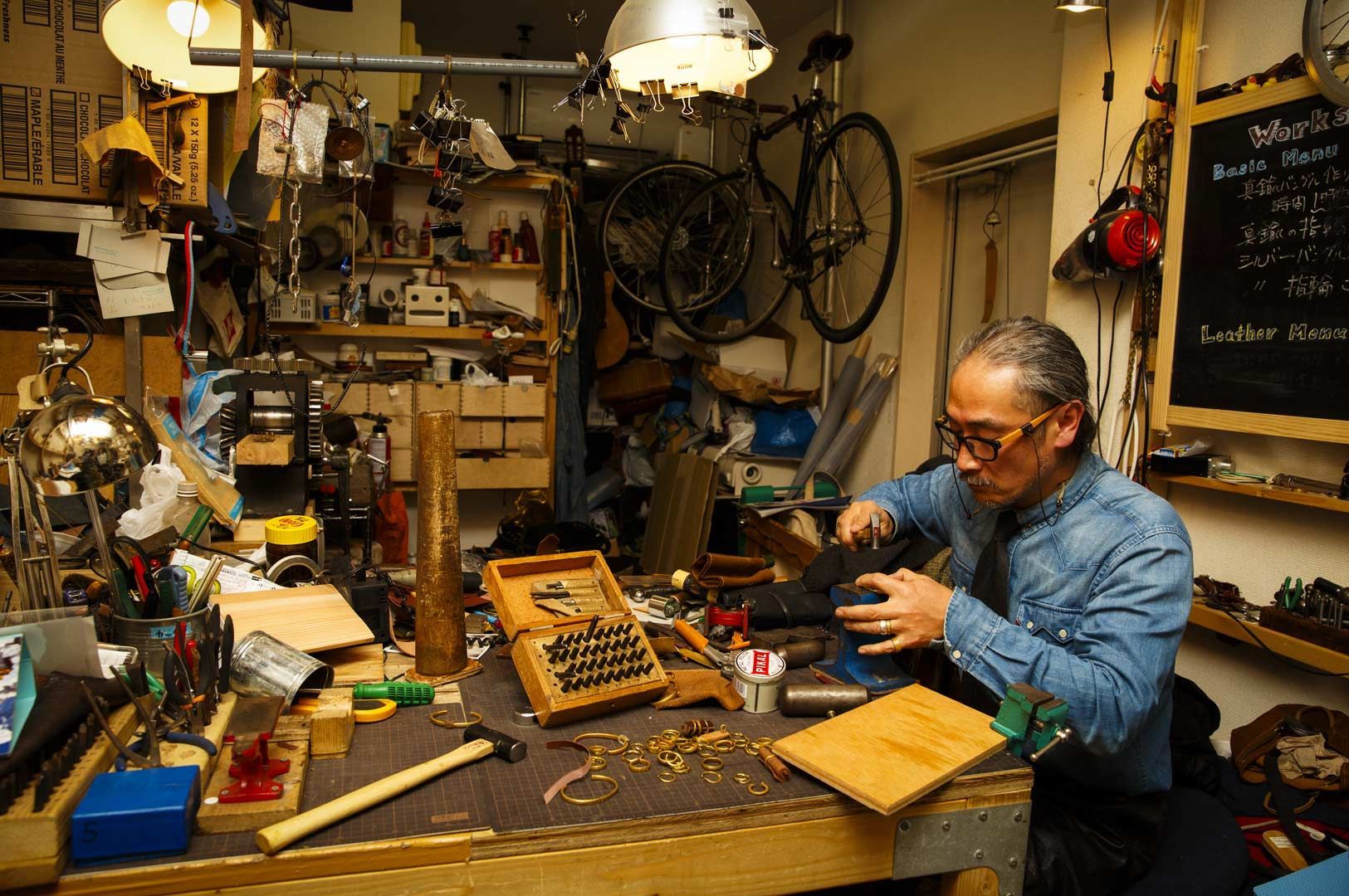 Craftsperson working at a cluttered workshop table filled with tools, leatherworking supplies, and equipment.