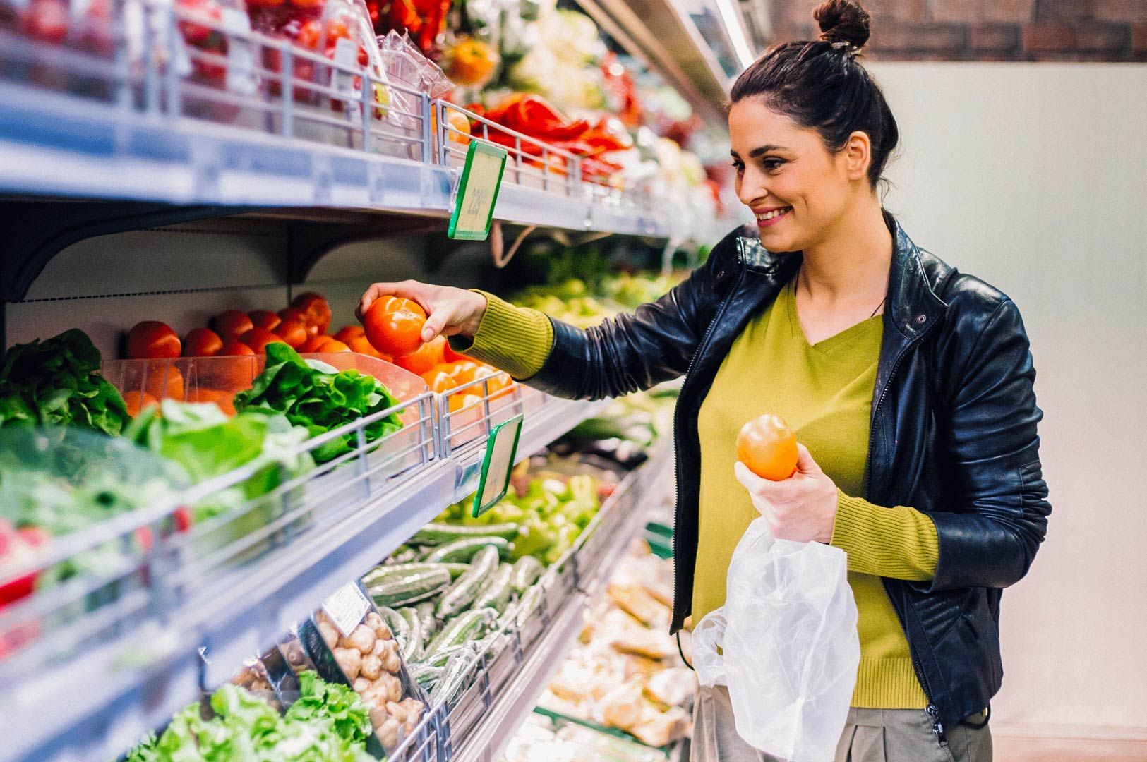 Person choosing fresh tomatoes from the produce section of a supermarket while holding a reusable bag.