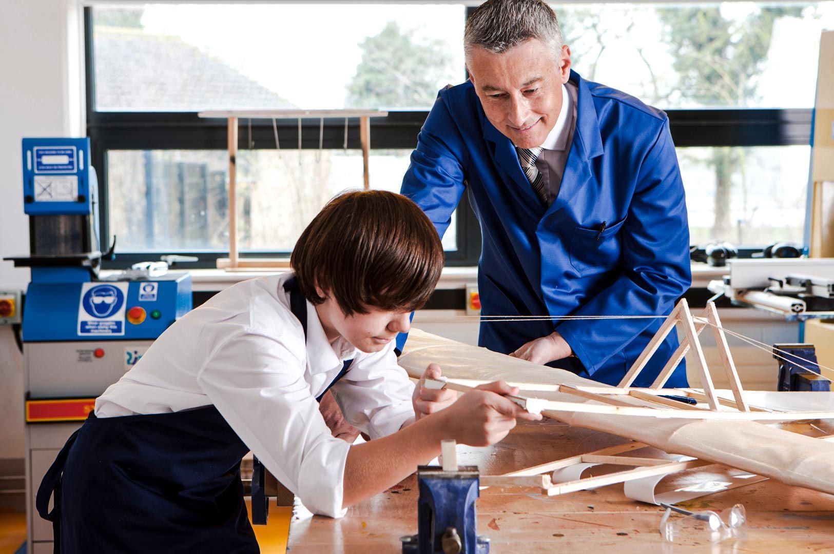 A workshop setting where an instructor guides a learner working on a wooden model structure using tools on a workbench.