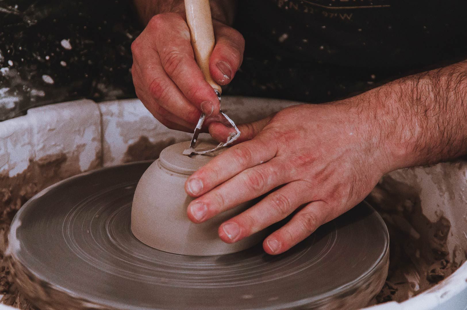Close-up of hands shaping clay bowl on pottery wheel with carving tool, fingers covered in clay.