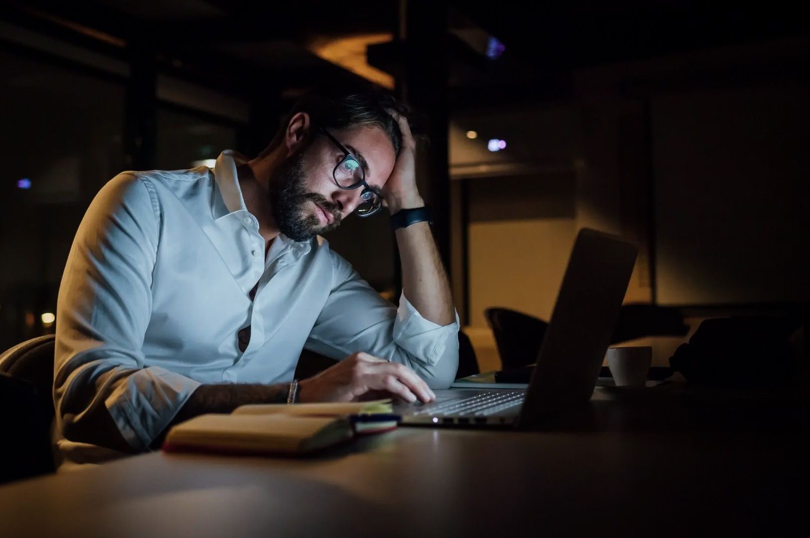 Individual sitting at a desk with a laptop, notebook, and coffee cup in a dimly lit workspace.