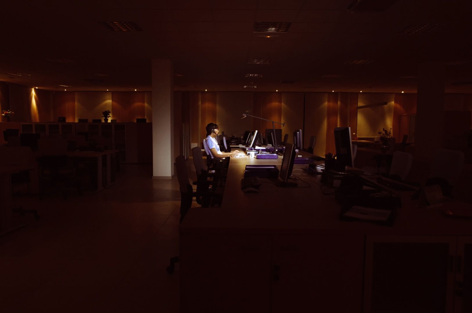 Person sitting at a desk in a dark office illuminated by computer screens, surrounded by rows of empty workstations.