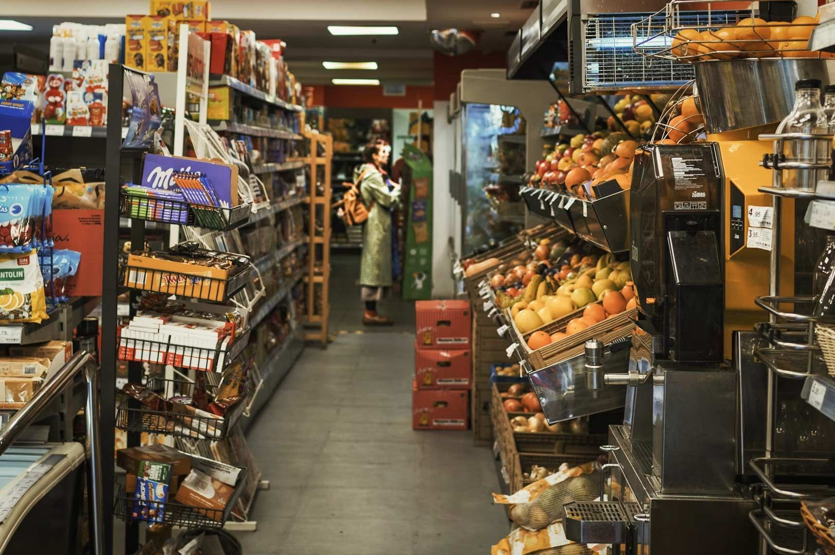Inside a local grocery store with shelves stocked with snacks, produce, and packaged goods along a narrow aisle.