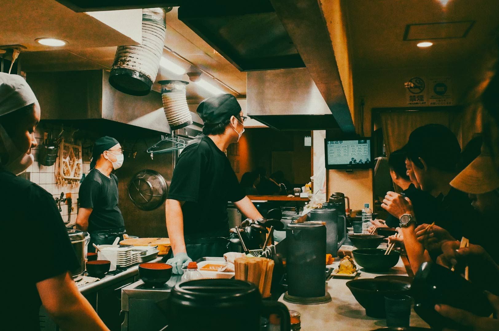 A busy restaurant kitchen with several people preparing food, surrounded by bowls, utensils, and cooking equipment, while diners eat at a counter nearby.