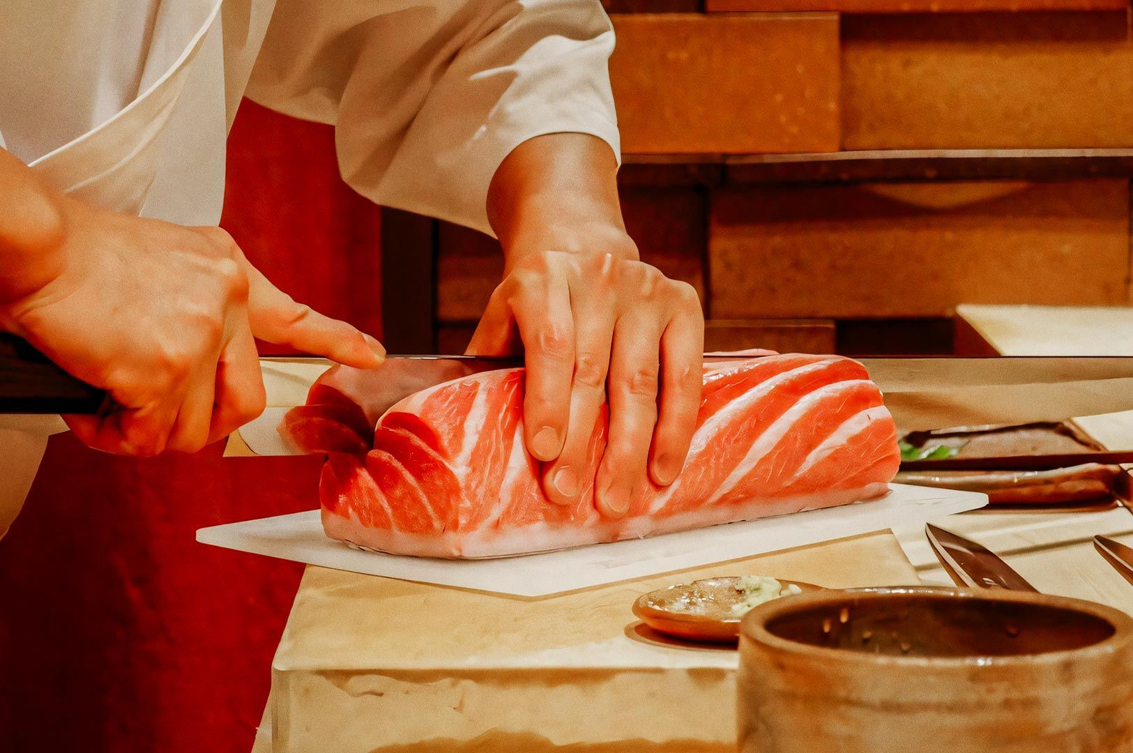 A chef slicing a thick cut of marbled raw tuna on a cutting board, surrounded by knives and small ceramic bowls in a sushi preparation setting.
