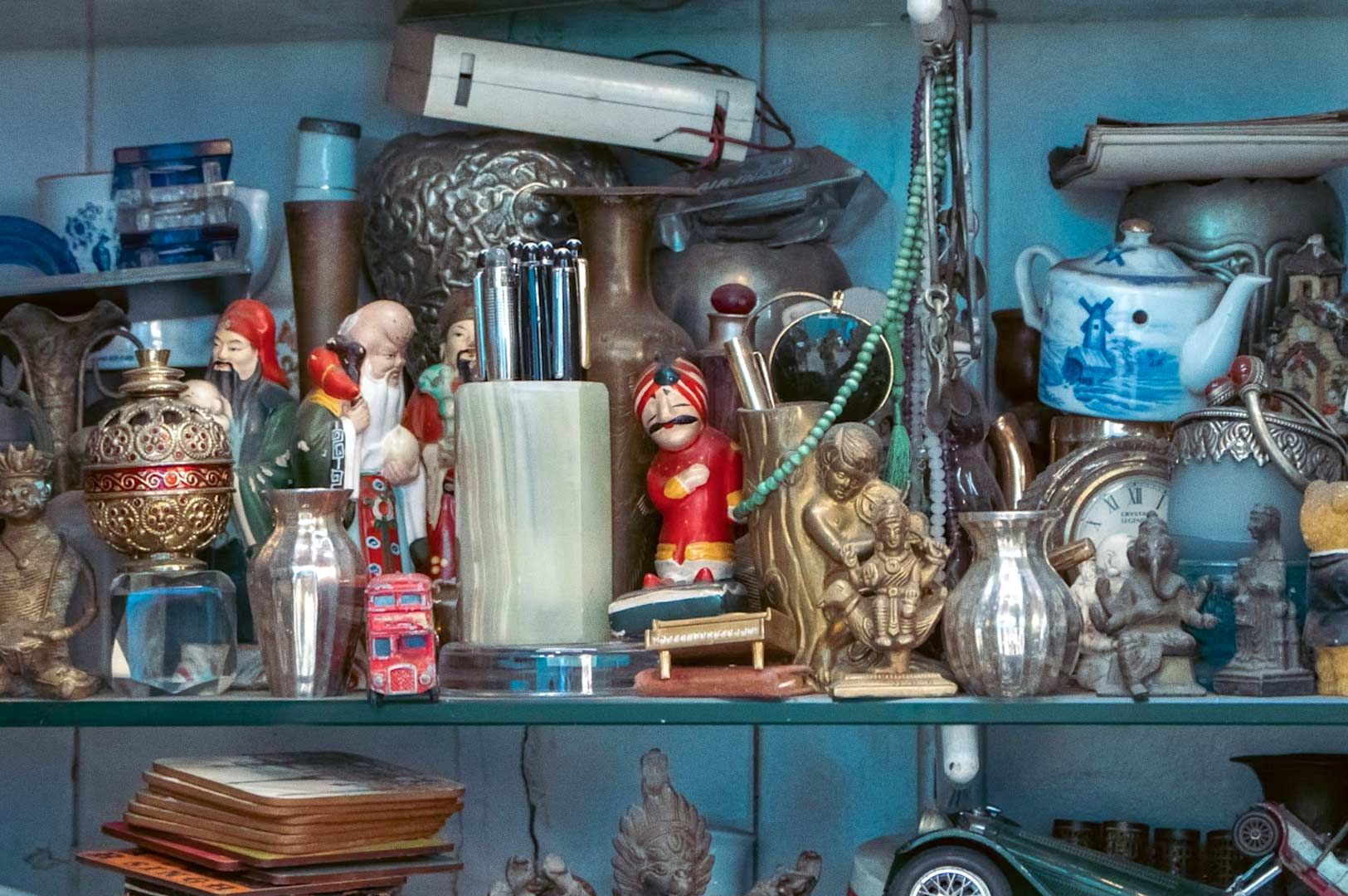 Cluttered decorative shelving showcasing cultural collectibles: figurines, ornate containers, a toy bus, vintage clock, and stacked coasters.