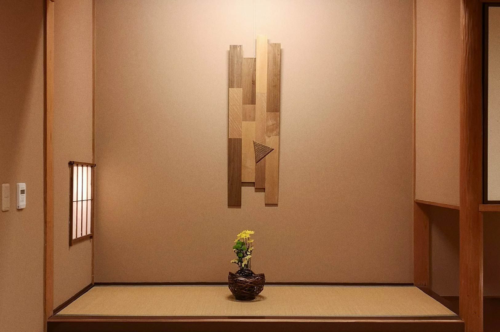 Minimalist Japanese tokonoma alcove featuring a wooden abstract wall art piece and a small flower arrangement in a woven vase, set against soft beige walls and tatami flooring.
