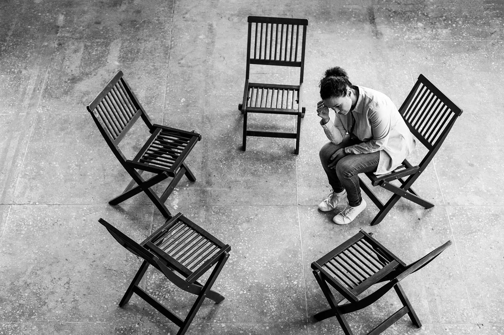 Solitary person seated on a wooden chair in the center of a circle of six empty wooden chairs, head bowed in a contemplative pose.