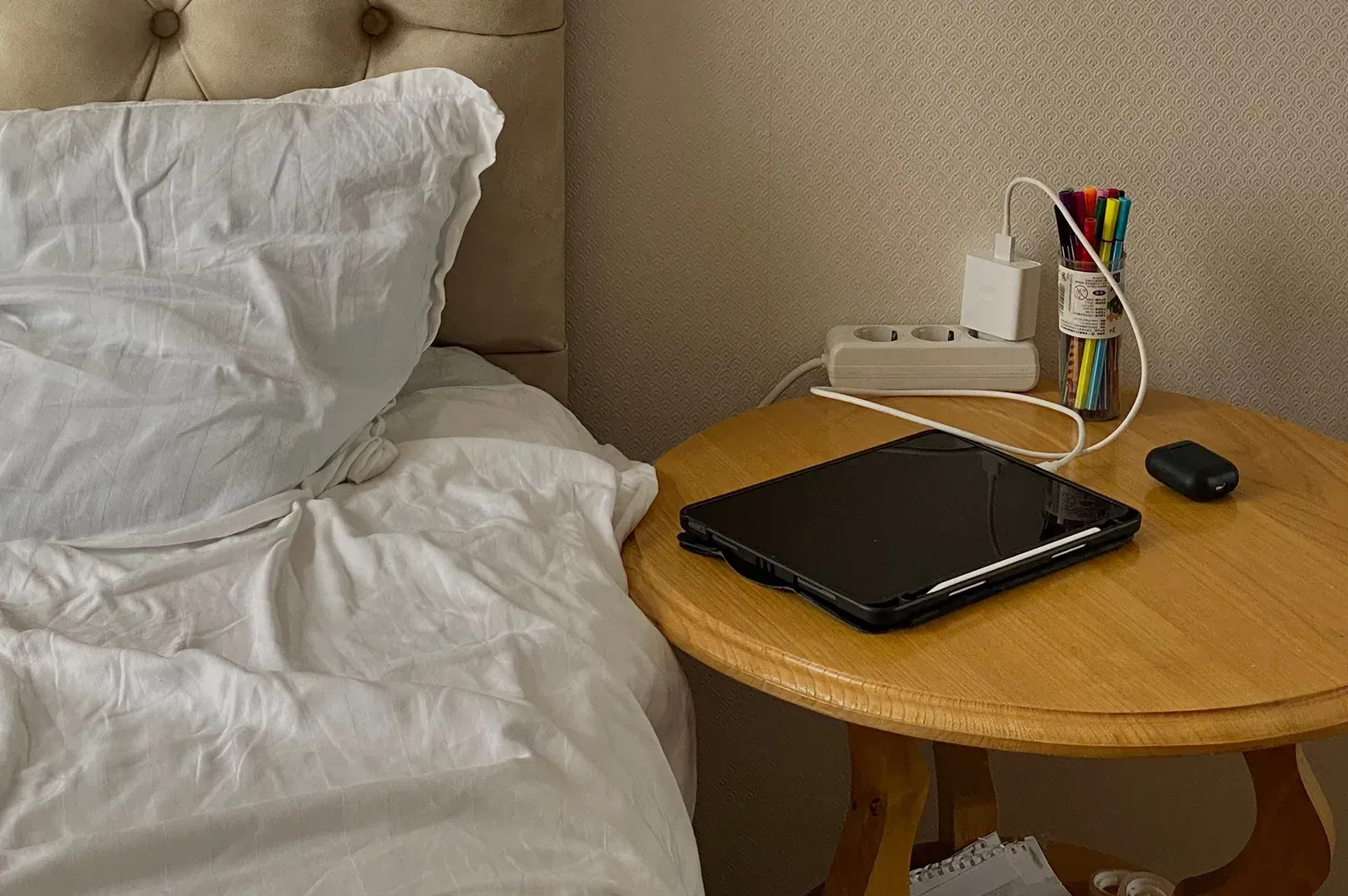 A quiet bedside table with a tablet, charging cords, and a cup of pens, depicting the maintenance of personal space and energy as a consistent, daily habit of self-respect.