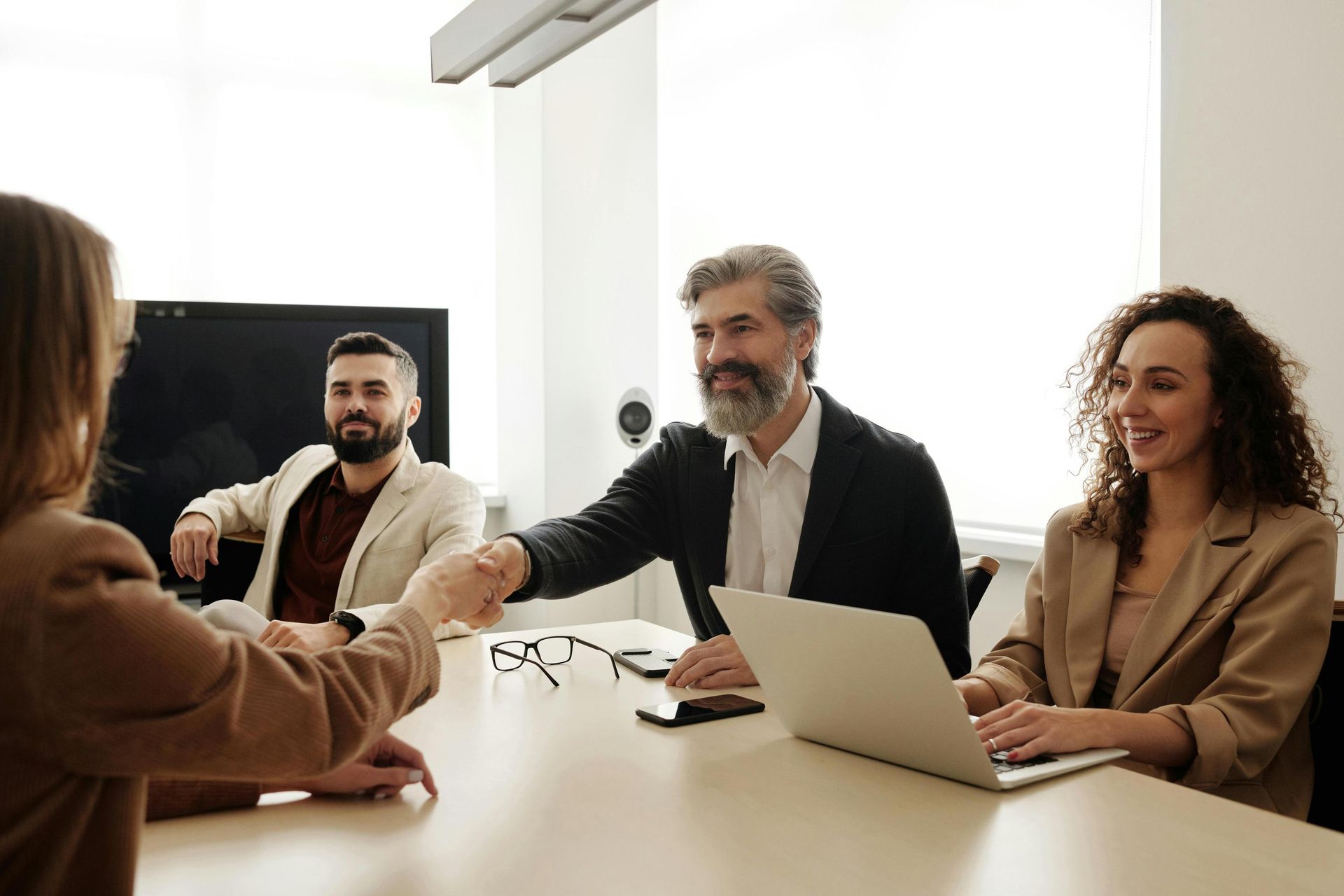 In a brightly lit office setting, a middle-aged man with a grey beard smiles as he shakes hands with a woman across a conference table. Their colleagues look on with pleasant expressions, creating a professional and welcoming atmosphere for the meeting.