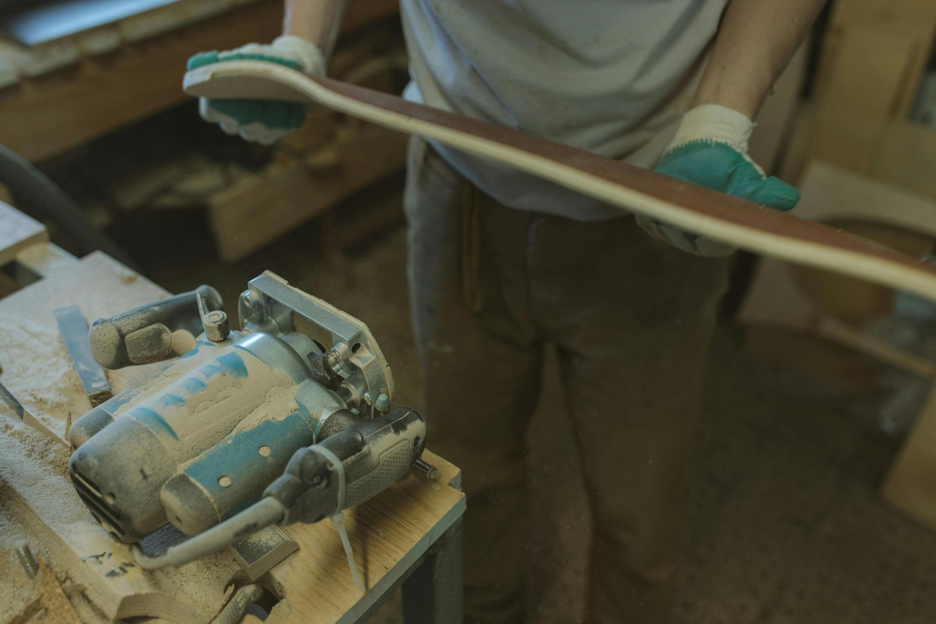 In a woodworking shop, a person wearing protective gloves holds a long, curved piece of wood that appears to be part of a skateboard deck. A dust-covered power router sits on a nearby workbench, surrounded by wood shavings and tools from the crafting process.