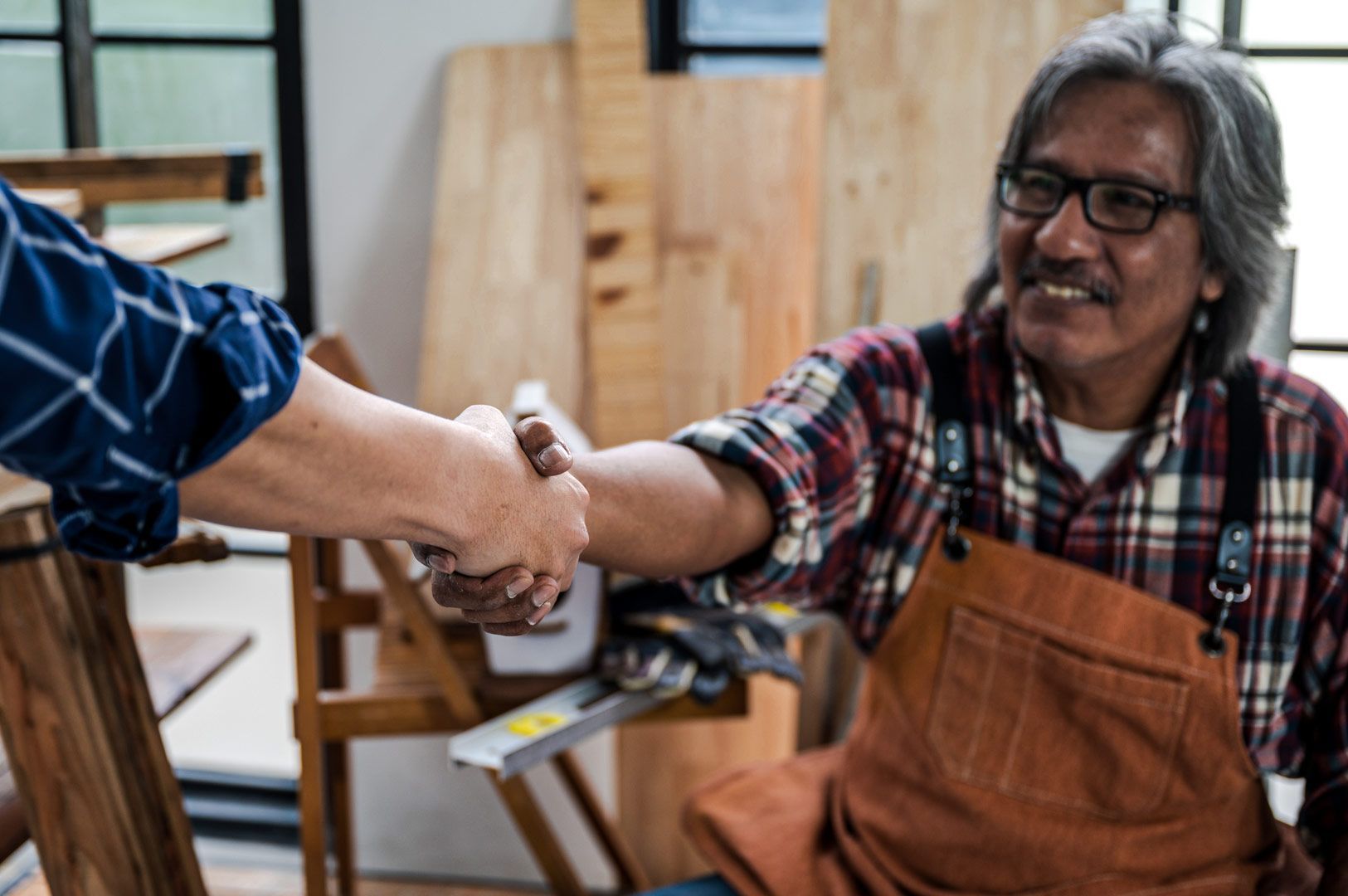 A woodworker in a workshop wearing a plaid shirt and apron shaking hands with another person, surrounded by wooden boards and carpentry tools.