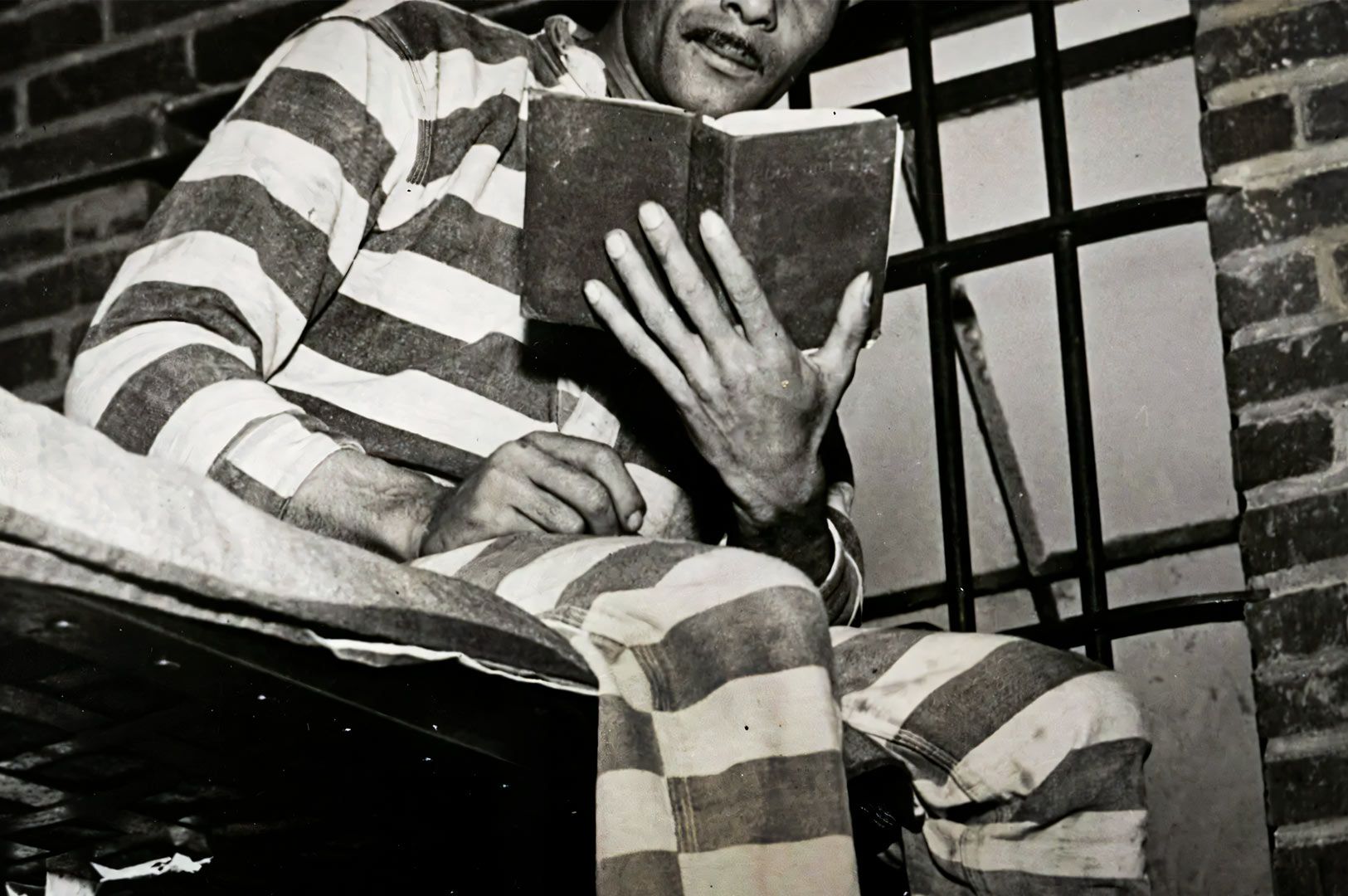 erson wearing striped prison clothing sitting on a bunk in a jail cell while reading a book, with metal bars and brick walls visible in the background.