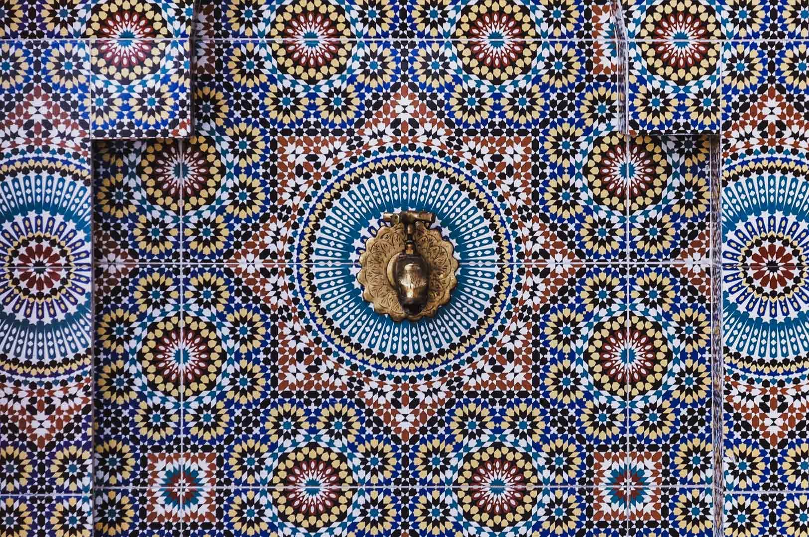 Moroccan zellige tile wall with colorful geometric starburst patterns and ornate brass hand-shaped door knocker centerpiece.