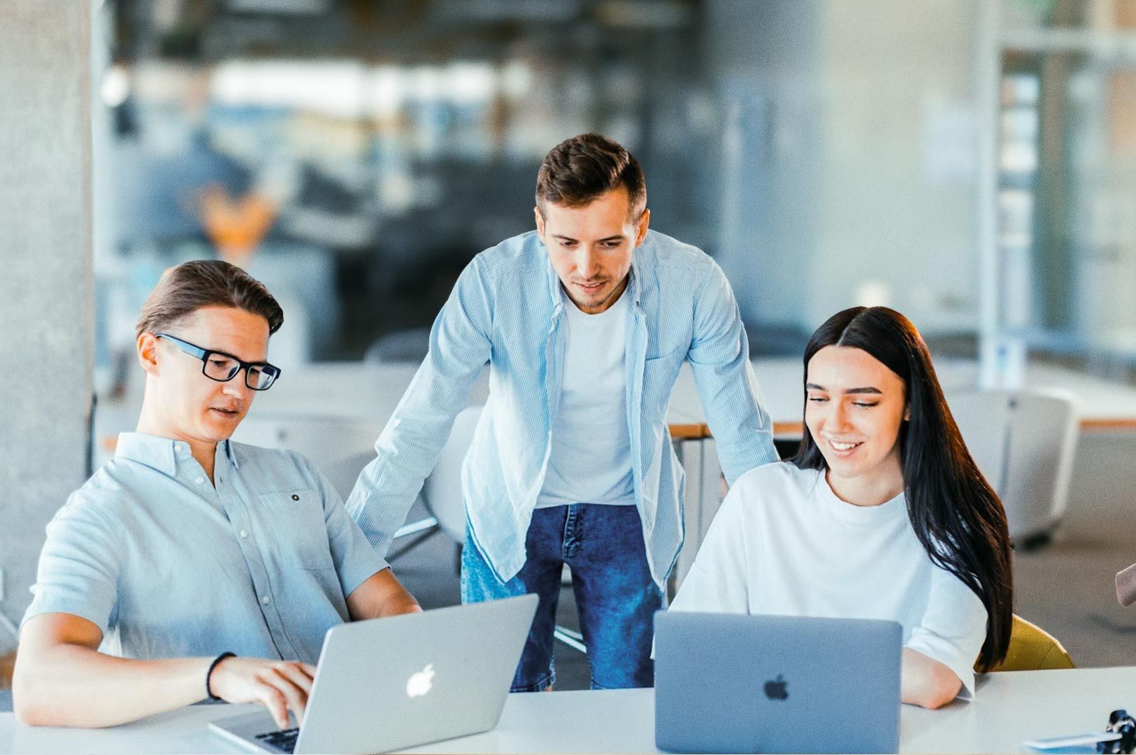 Group of colleagues collaborating around laptops in a modern open office environment, discussing a project at a shared workspace.