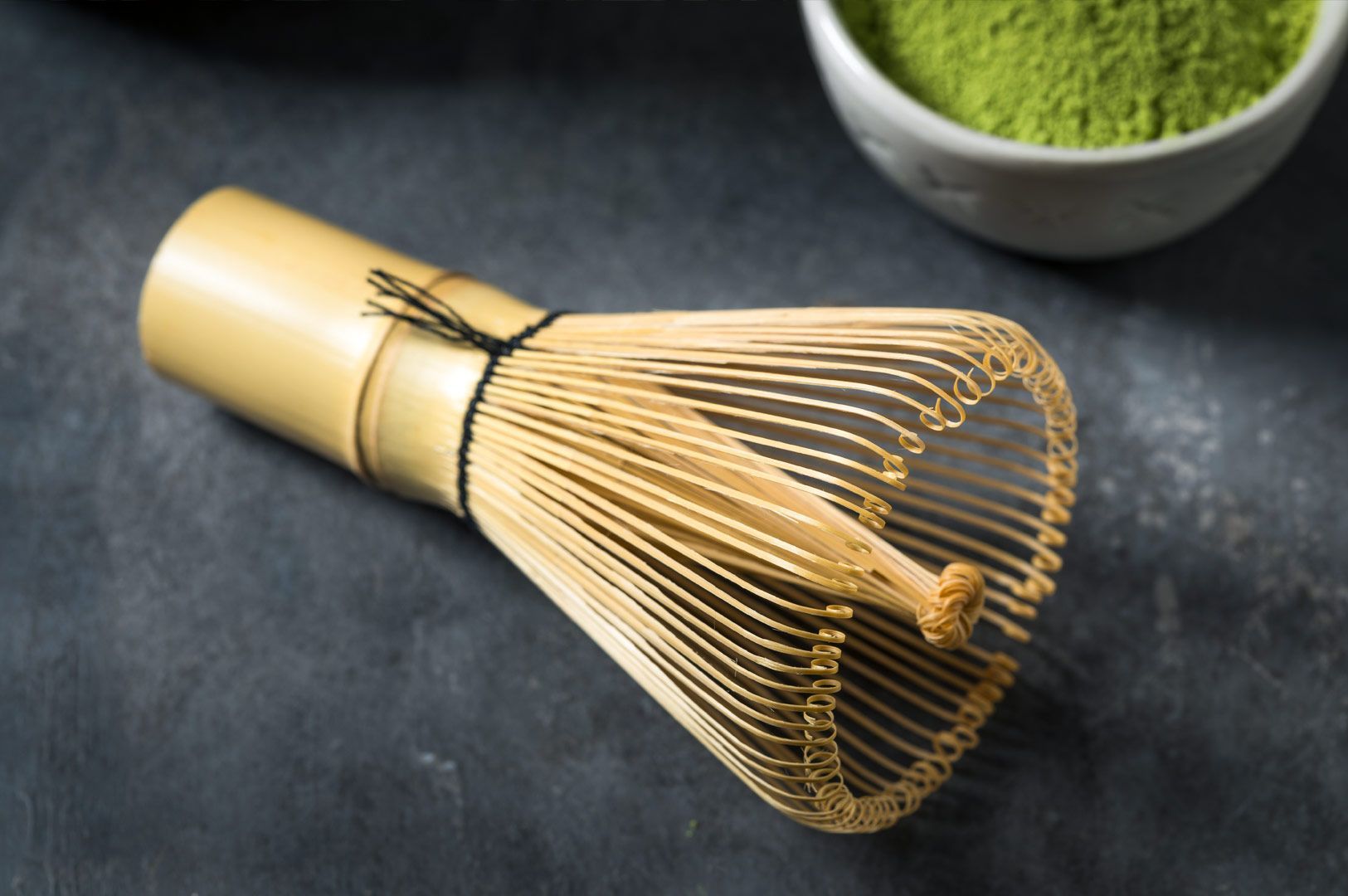 Traditional bamboo matcha whisk placed on a dark surface with a bowl of green matcha powder in the background.