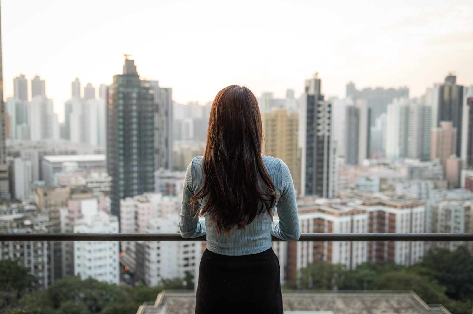 Individual standing on a high balcony observing a panoramic city skyline with tall residential and commercial buildings.