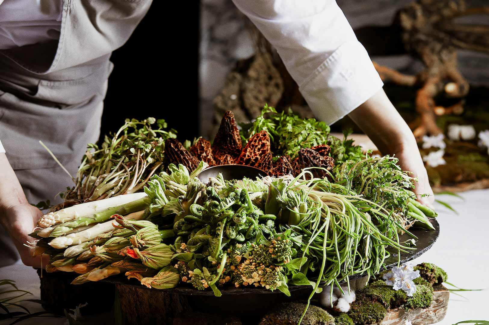 A large platter of fresh spring vegetables, including fiddlehead ferns, asparagus, greens, and edible flowers, arranged decoratively while a chef places the dish on a table.