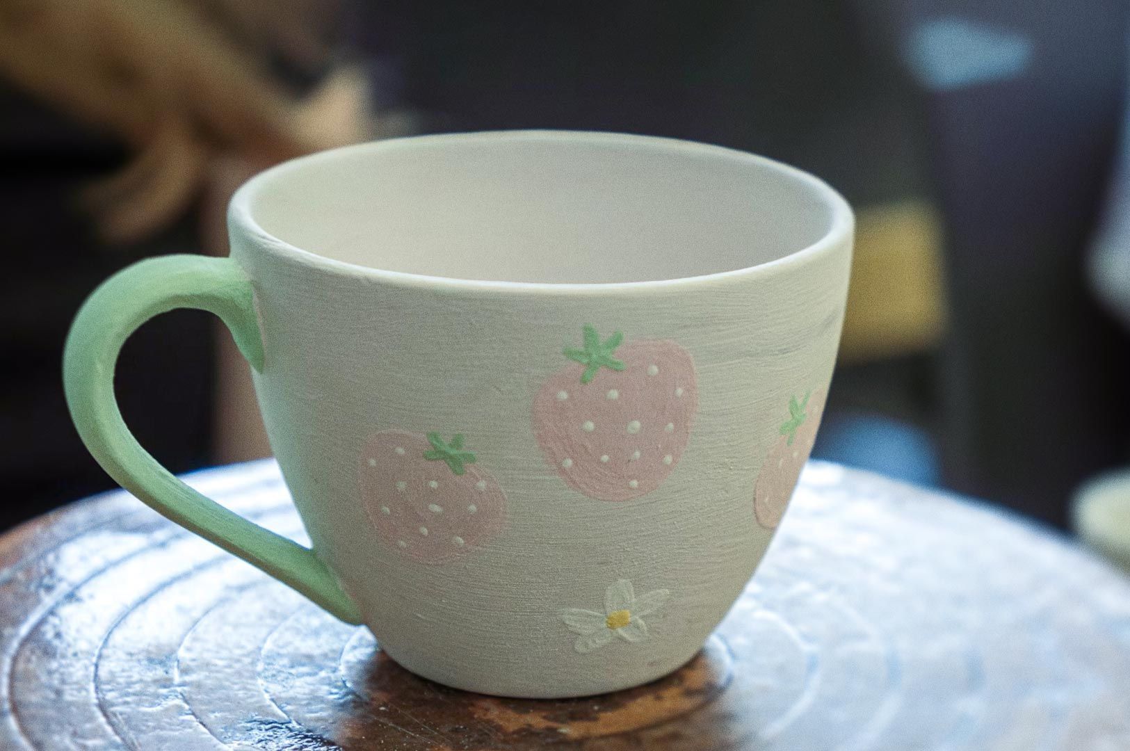 Handmade ceramic mug with pastel green handle and hand-painted pink strawberries on a light beige body, photographed on a textured wooden pottery wheel in a studio.