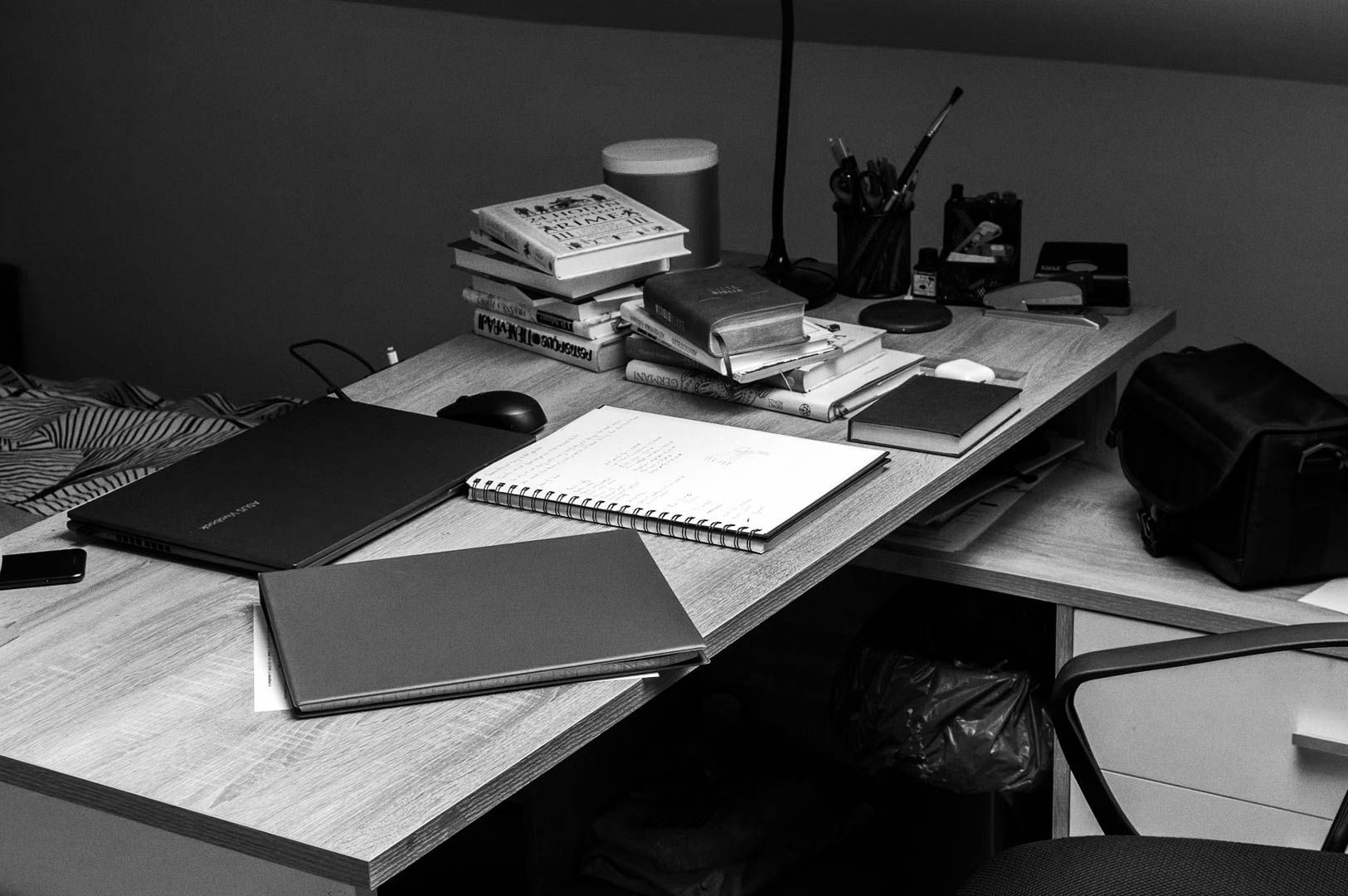 Study desk with an open spiral notebook of handwritten notes, closed laptop, mouse, stack of books, pen holder, small speaker, and camera bag arranged for focused work.