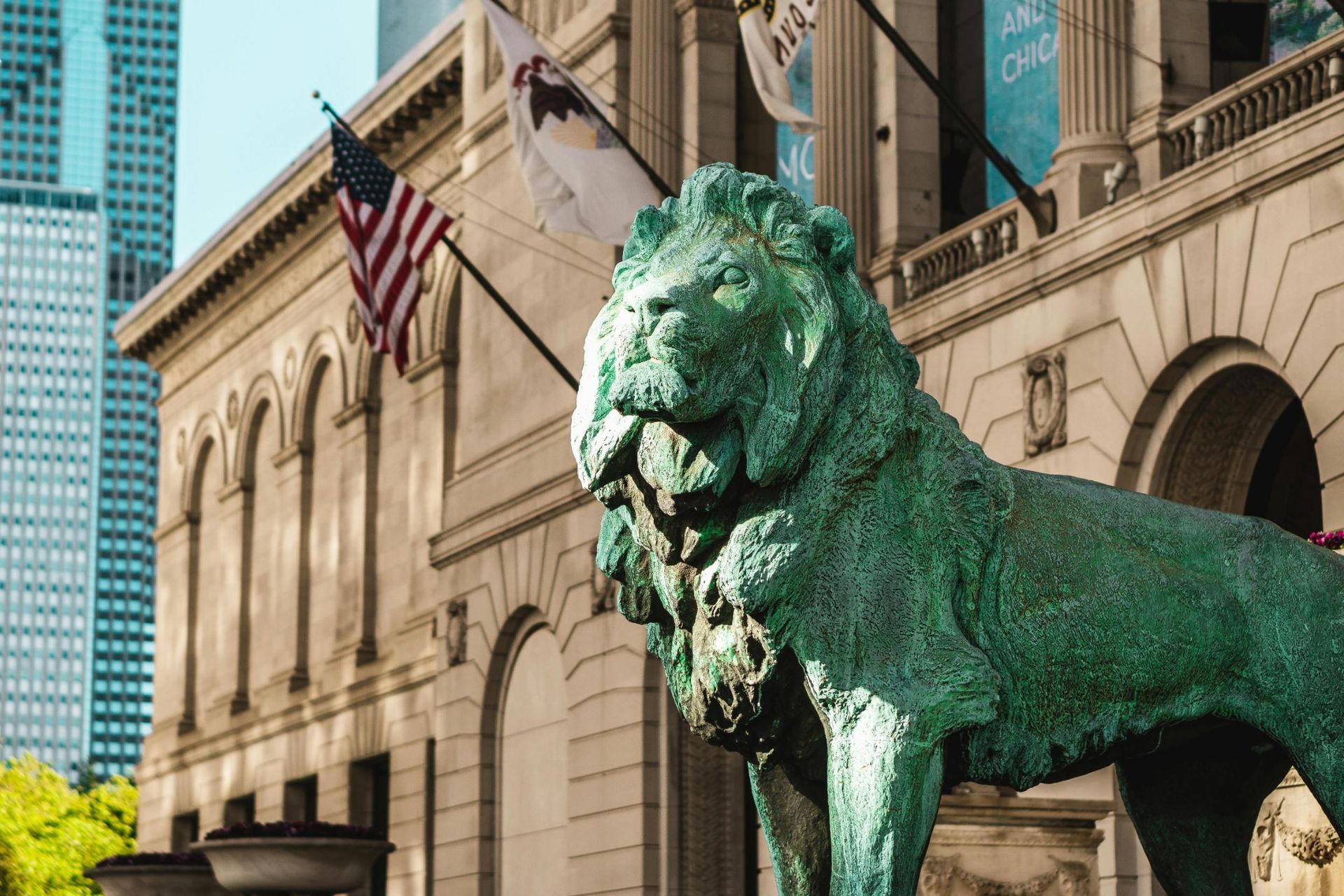 A weathered, green-patina lion statue stands prominently in the foreground against a backdrop of grand, neoclassical architecture adorned with the American flag. In the distance, a modern glass skyscraper rises above the historic stone building, highlighting a contrast between classic and contemporary urban design.